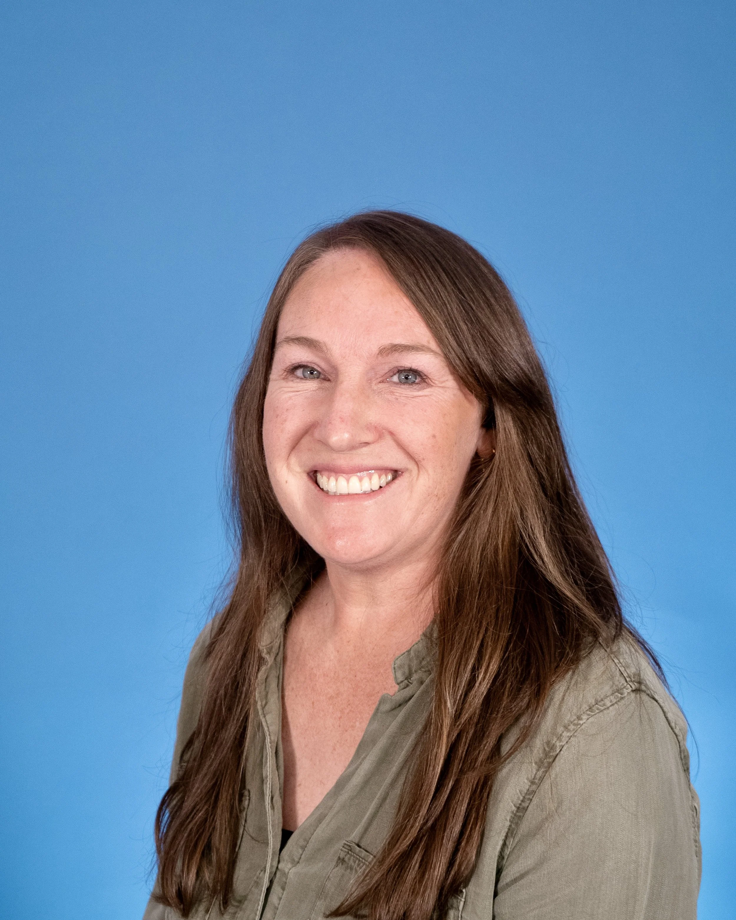 A woman with long brown hair and blue eyes smiling, wearing an olive green jacket against a blue background.