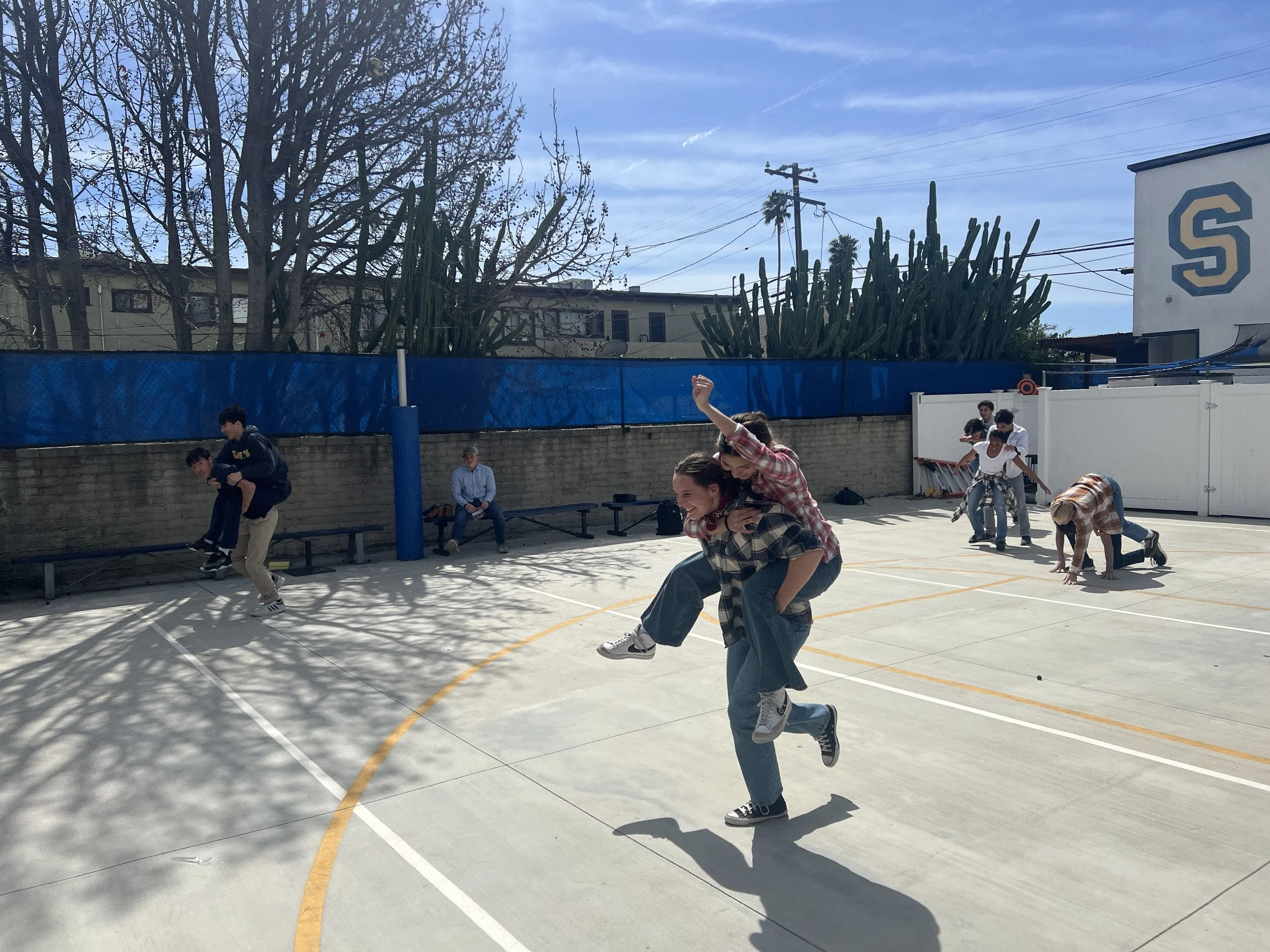 Young people playing football on an outdoor basketball court with trees, power lines, and a building with a large letter 'S' in the background on a sunny day.