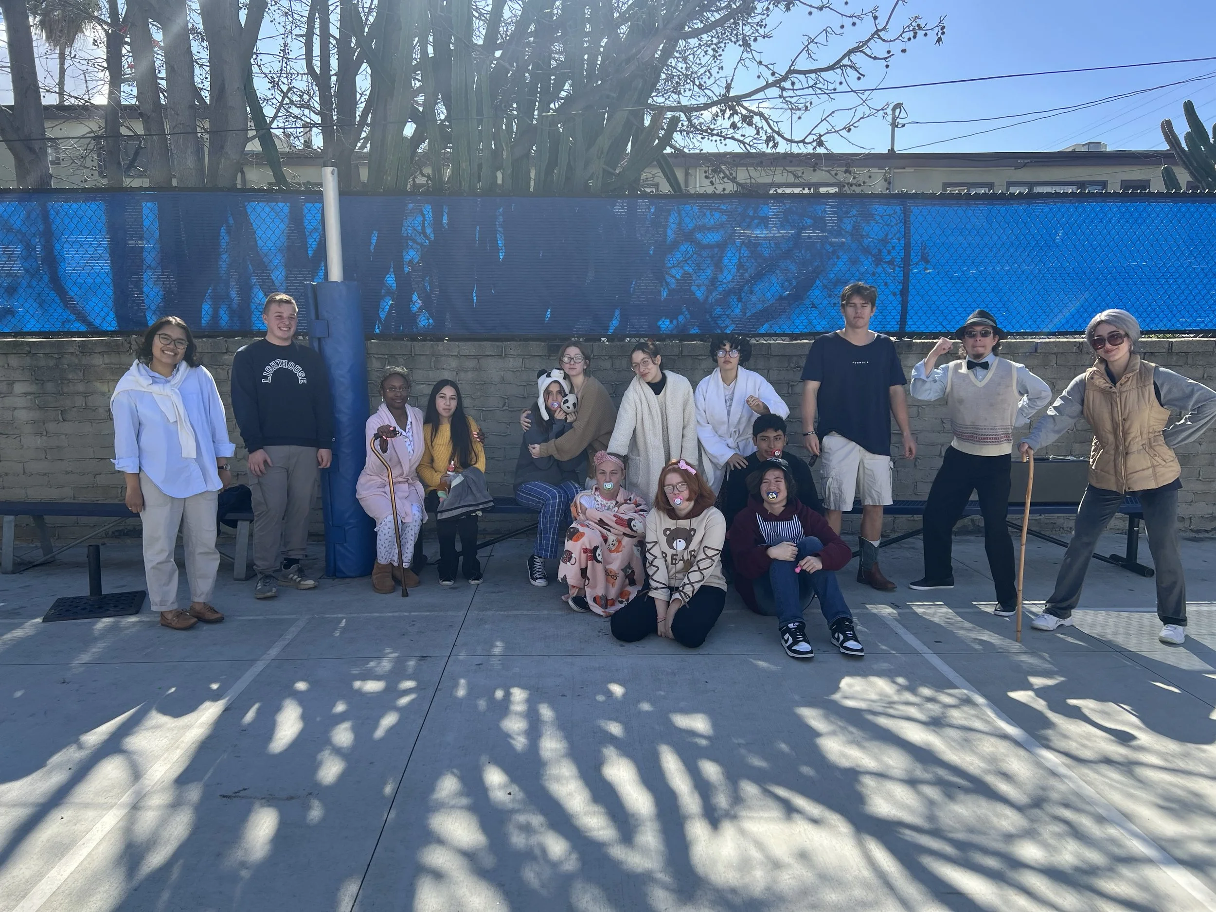 A group of children and teenagers dressed in costumes, posing for a photo outdoors in front of a brick wall with shadow patterns on the pavement, with trees, blue fence, and building in the background on a sunny day.
