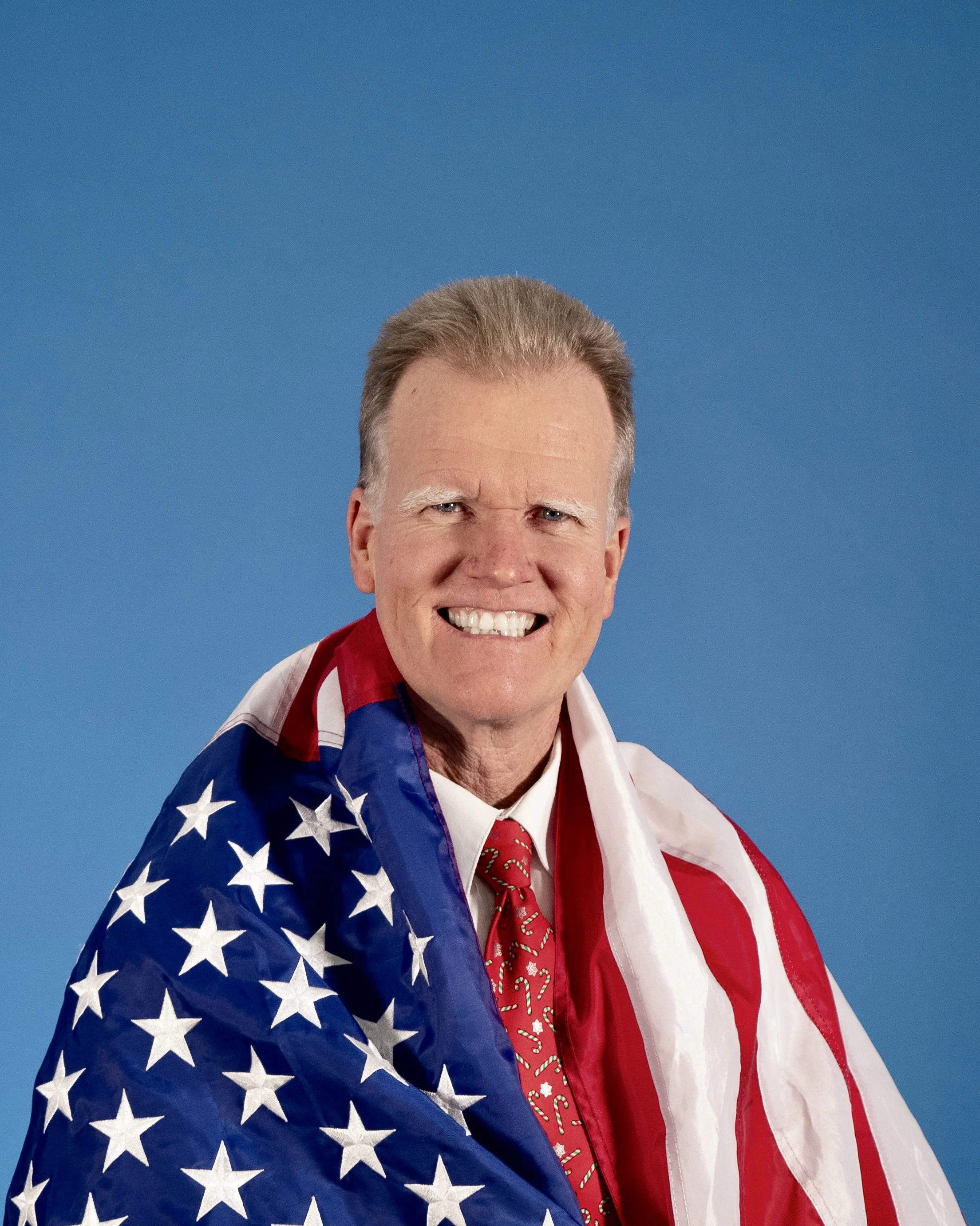 A smiling man with gray hair draped in an American flag against a blue background.