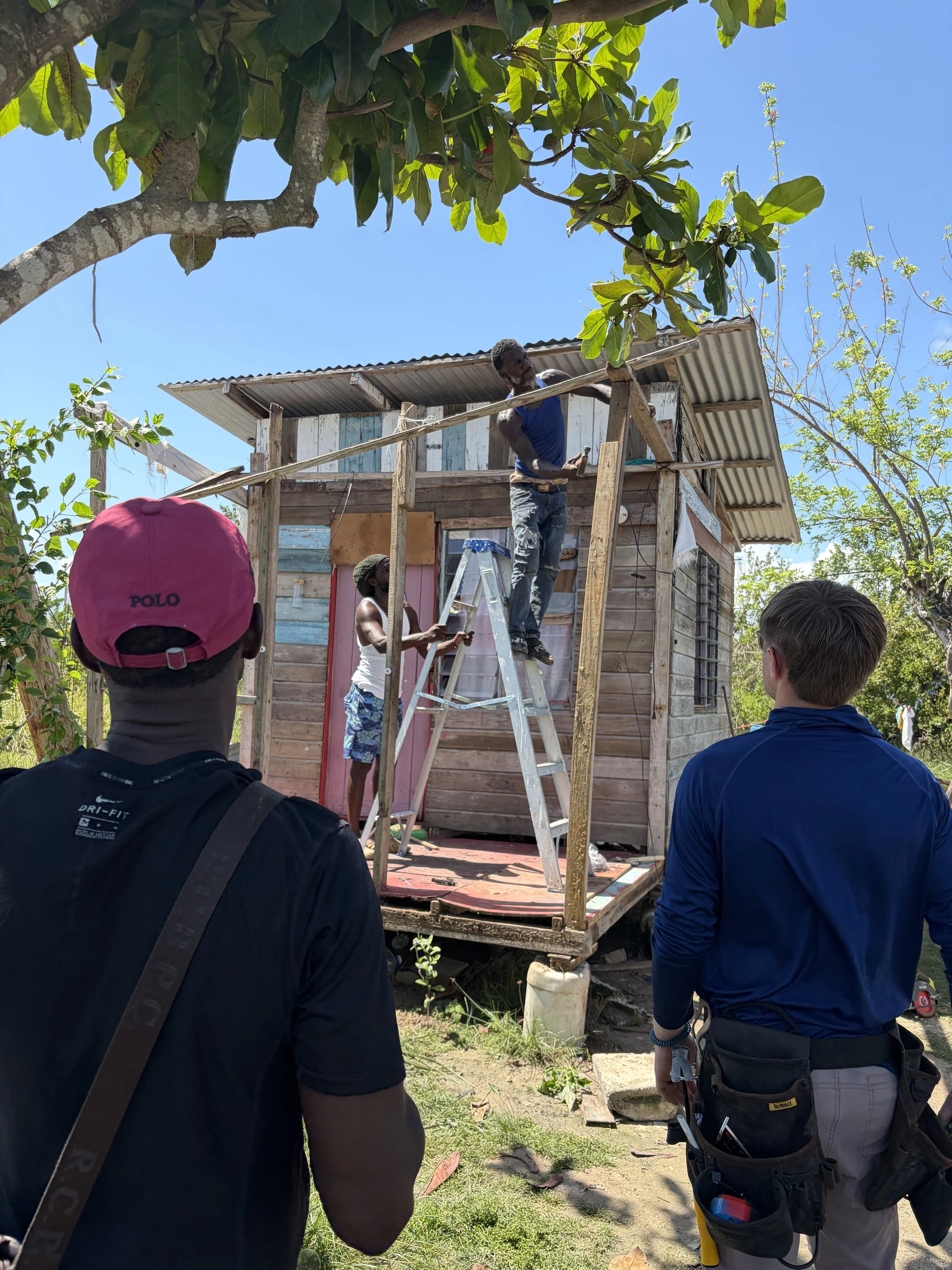 A group of people are gathered outdoors watching a man work on a wooden structure, possibly building or repairing it, with trees and a small house in the background and a clear blue sky.