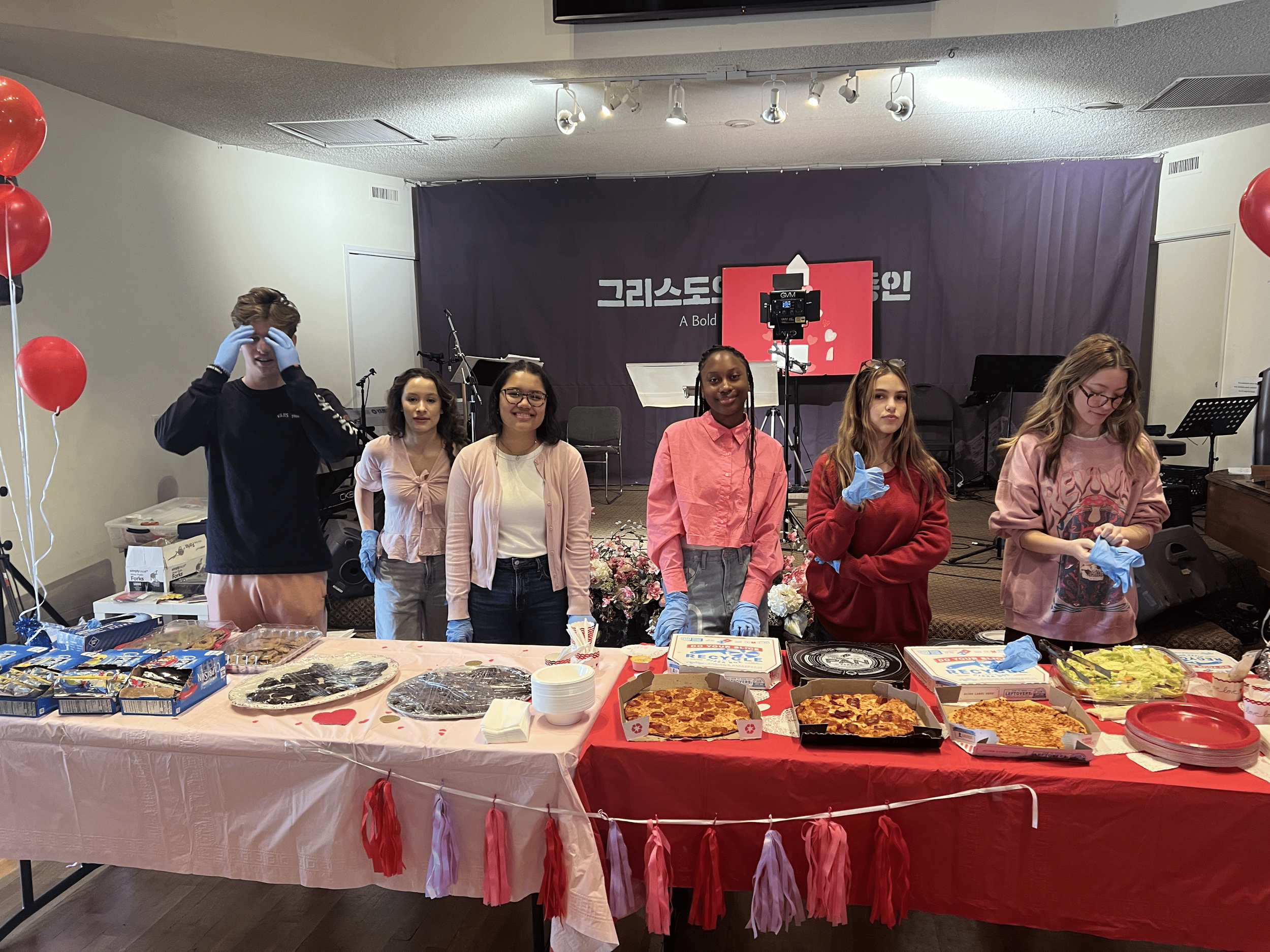 Group of young people standing behind a table with various food items, at an indoor event or celebration, with balloons on the side and a stage in the background.