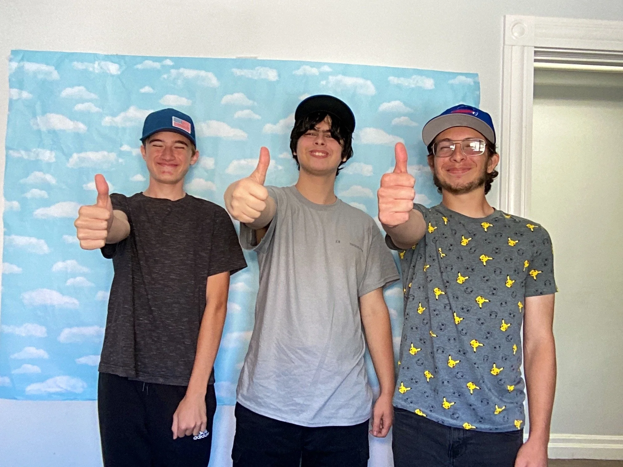 Three young men standing in front of a blue sky with clouds backdrop, giving thumbs up and smiling, wearing casual clothing and hats.