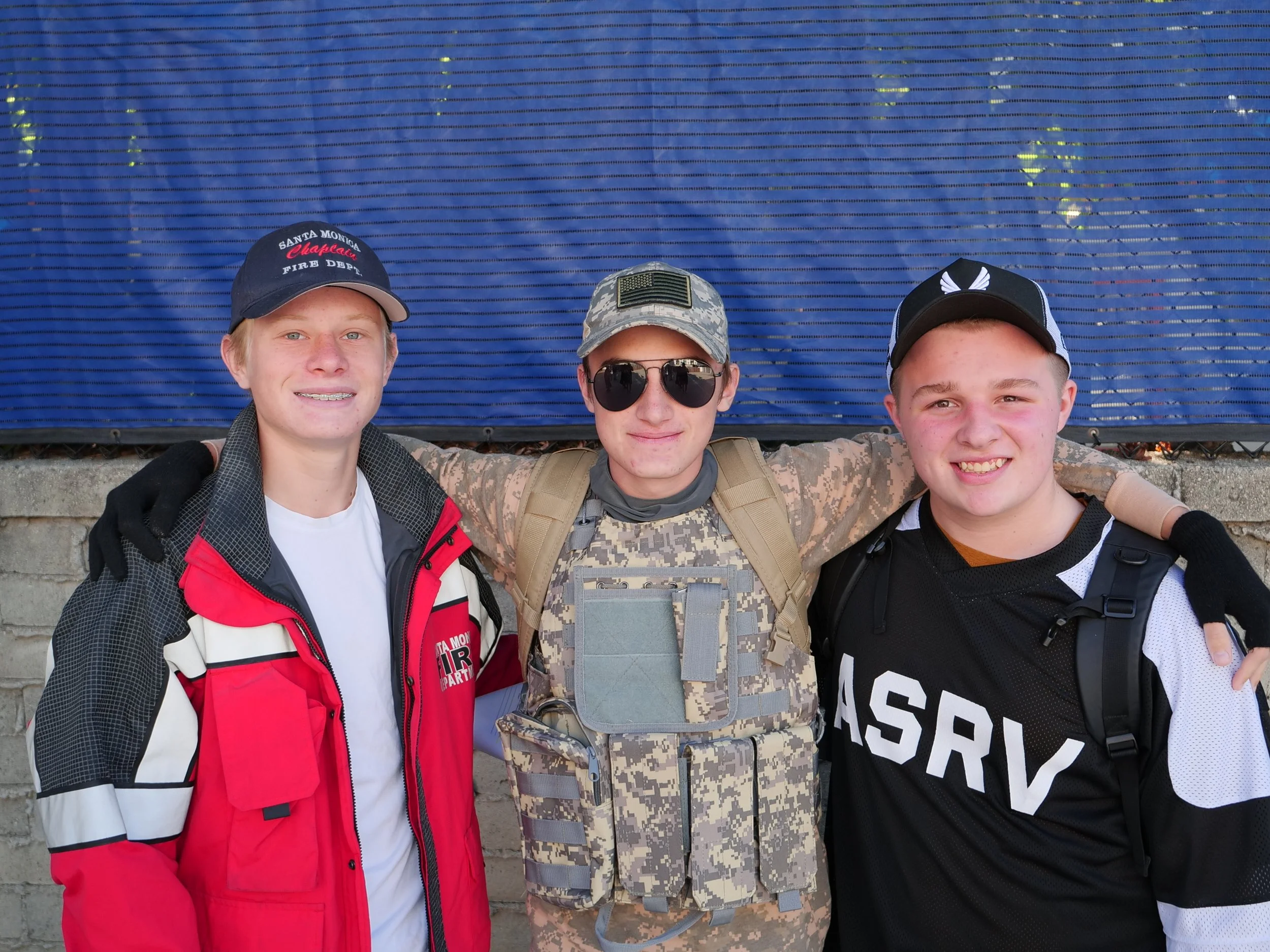 Three young men standing together outdoors, smiling with their arms around each other's shoulders. They are dressed in casual and military-style clothing, standing in front of a blue backdrop.