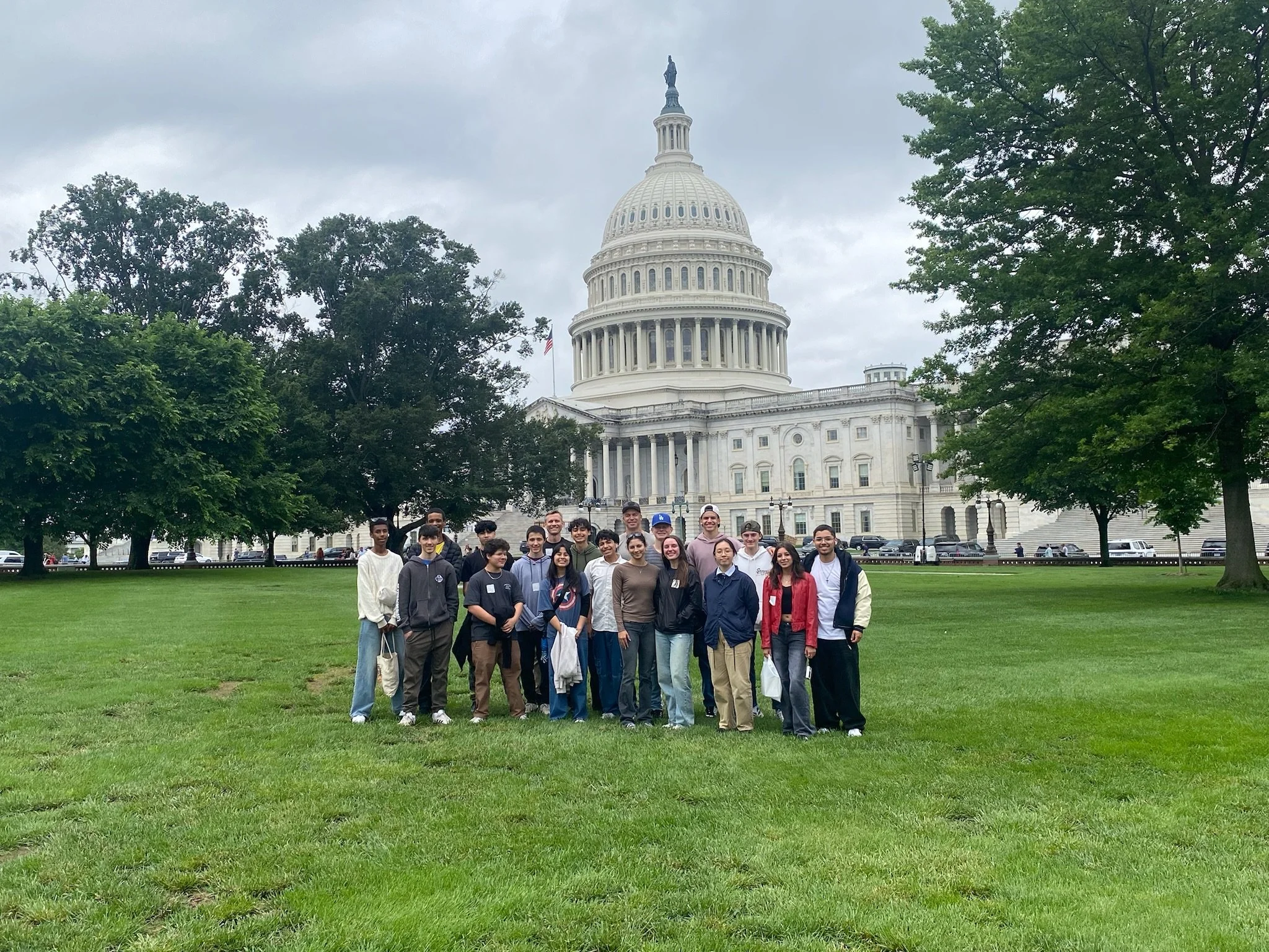A group of people standing on a grassy area in front of the U.S. Capitol building in Washington, D.C., under a cloudy sky with trees surrounding them.