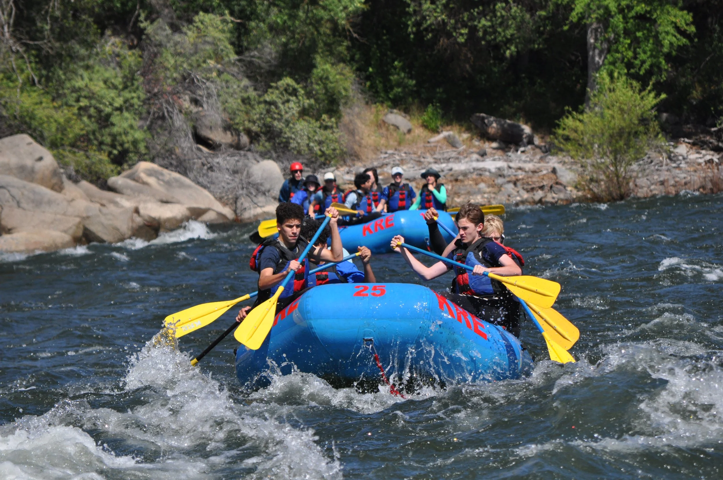 Group of people white water rafting on a river with rocks and trees in the background.