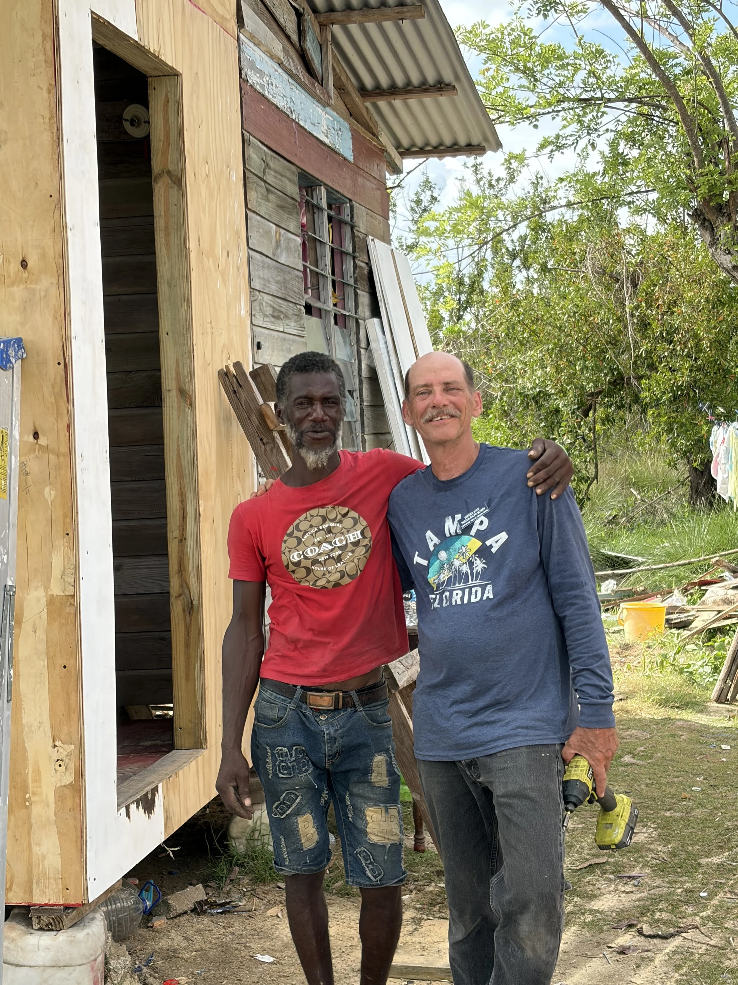 Two men standing outside a partially constructed wooden building, one holding a black and yellow power drill and the other wearing a red shirt, with trees in the background.