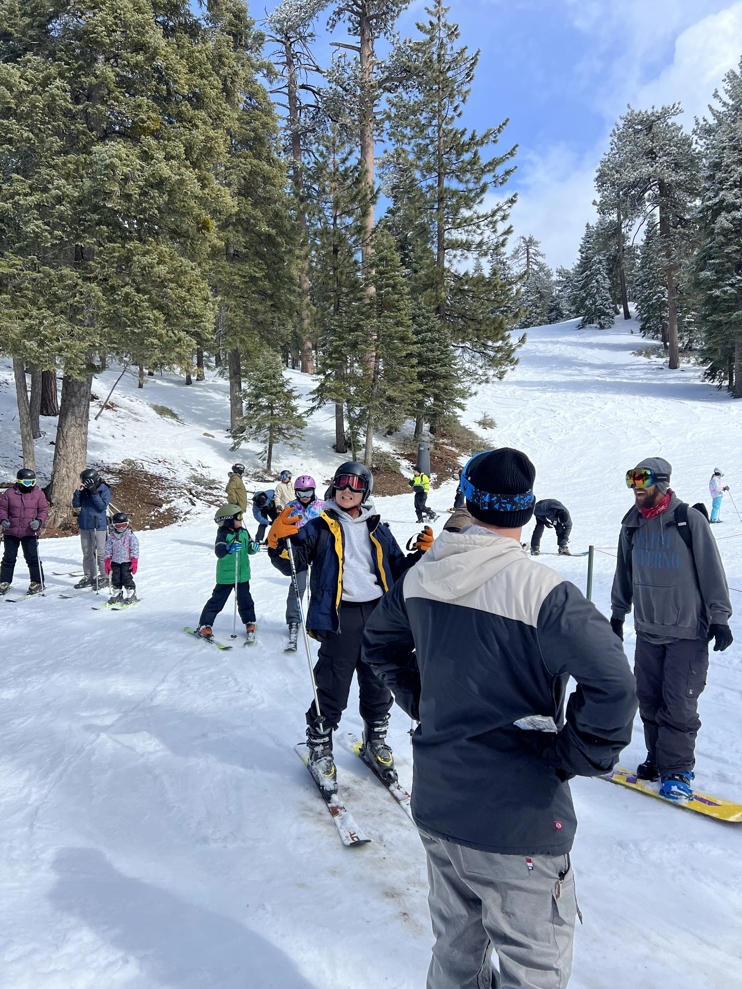 Group of people gathering at a snowy ski destination, some wearing helmets and ski gear, with a forested mountain backdrop and clear blue sky.