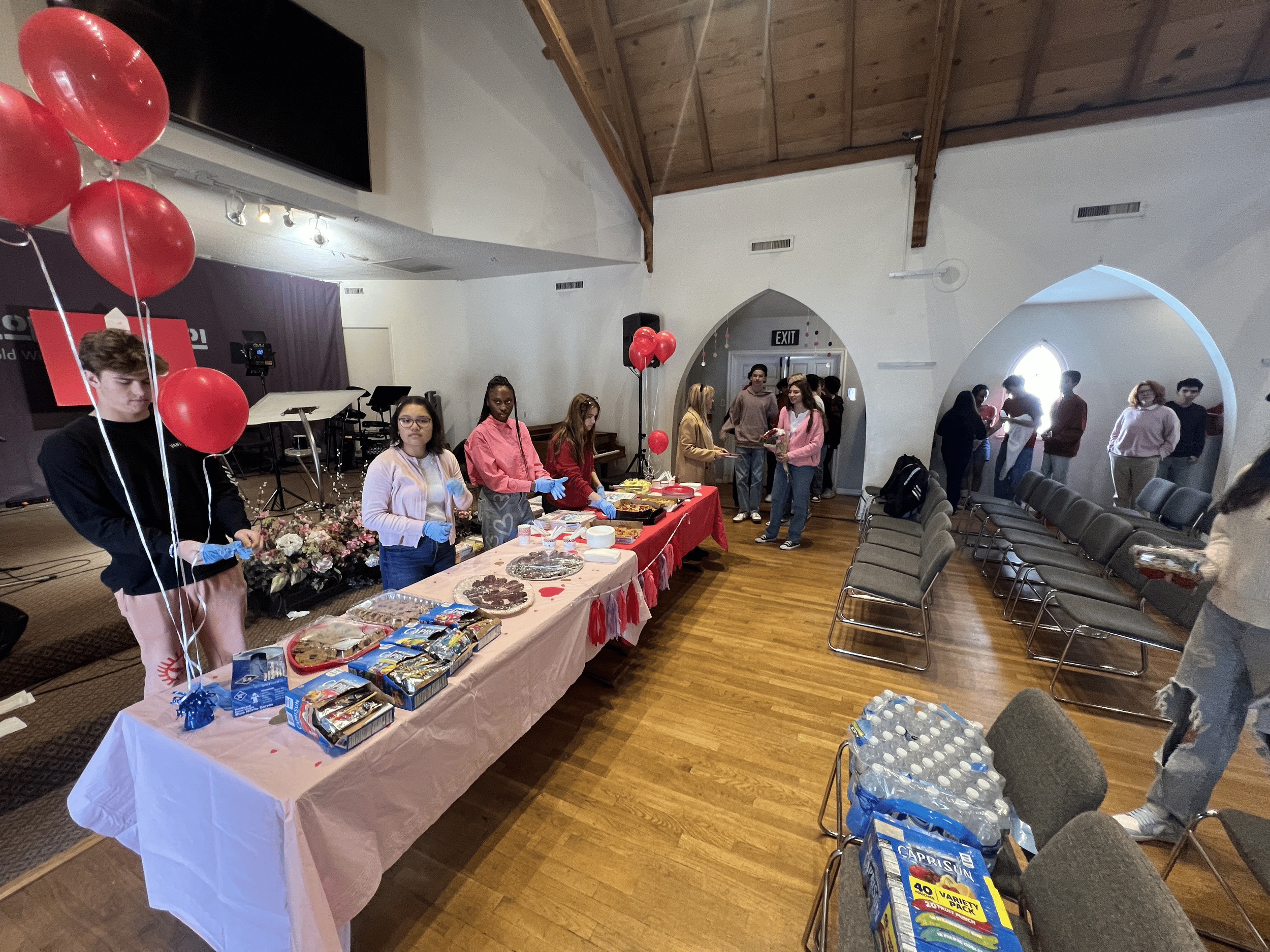 A group of people at a gathering in a room with a high wooden ceiling. Several individuals are standing behind a table with food, wearing gloves, and organizing snacks and treats. There are balloons, chairs, and more people mingling in the background