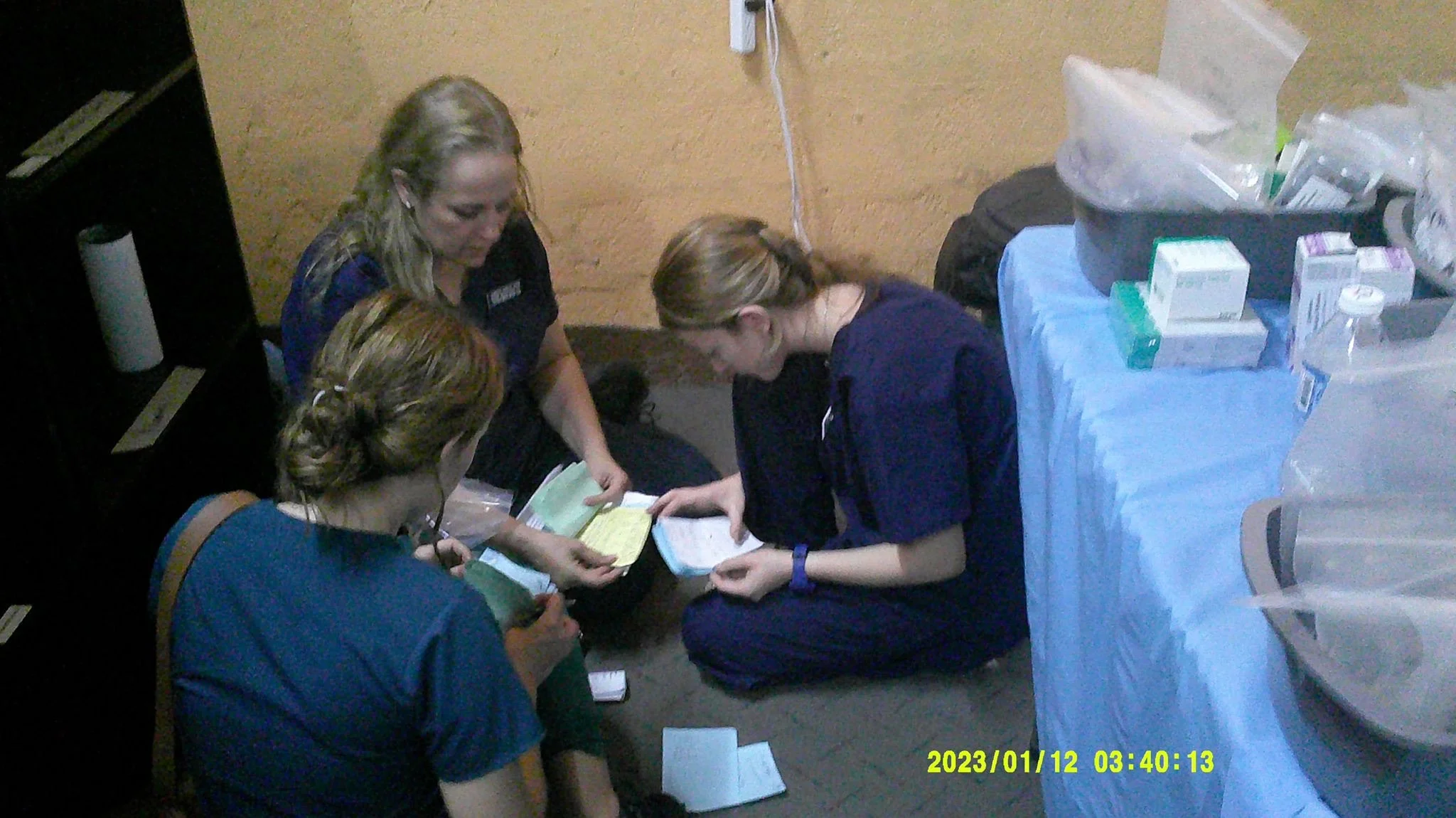 A group of five women, four in medical scrubs, are gathered around a table reviewing documents or medical records. There are medical supplies and boxes on the table beside them. The scene appears to be in a clinical or hospital setting, with a beige 