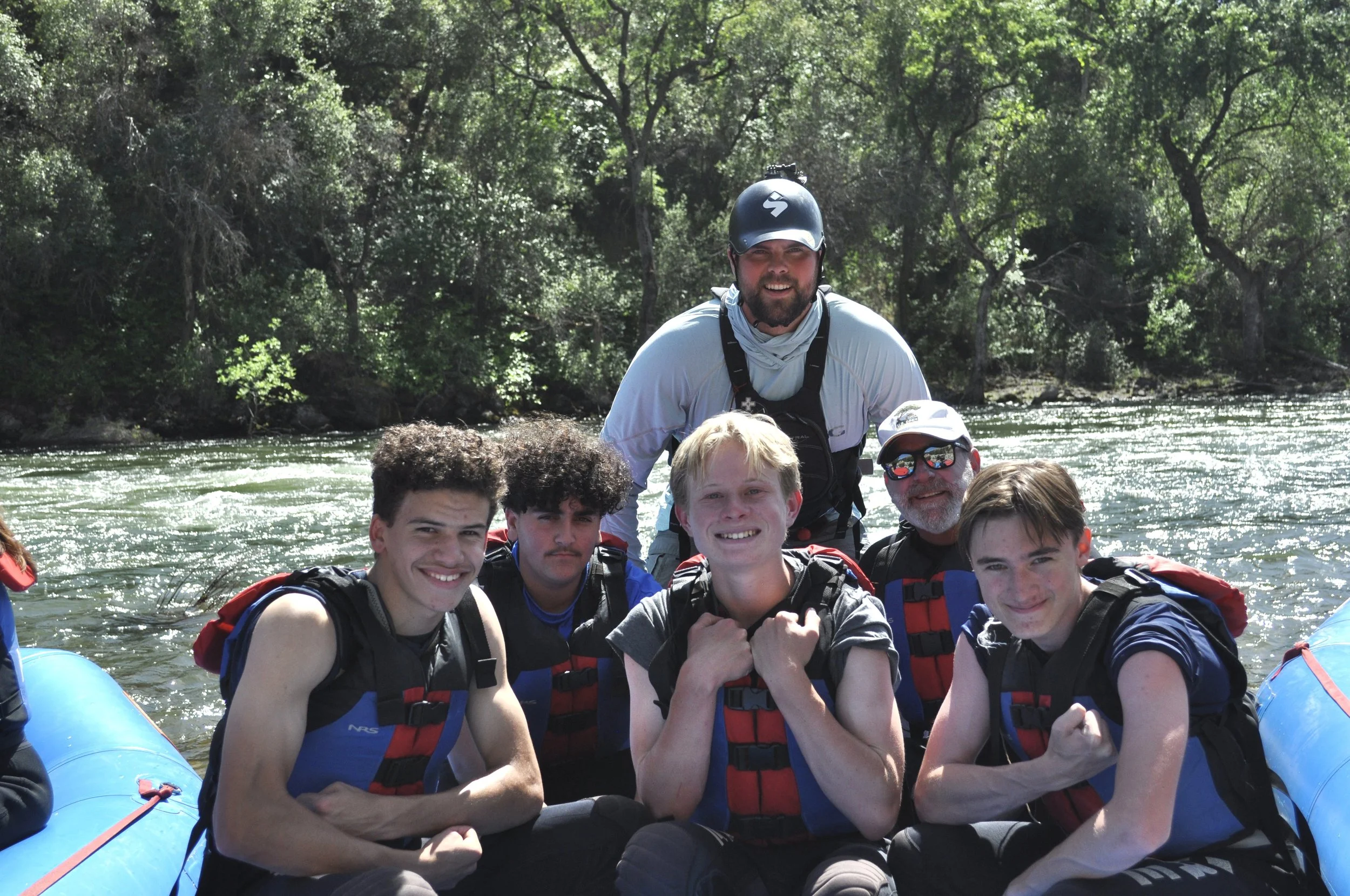 A group of young men and two adult men on a boat, wearing life jackets and helmets, smiling and posing for a photo on a river with trees in the background.