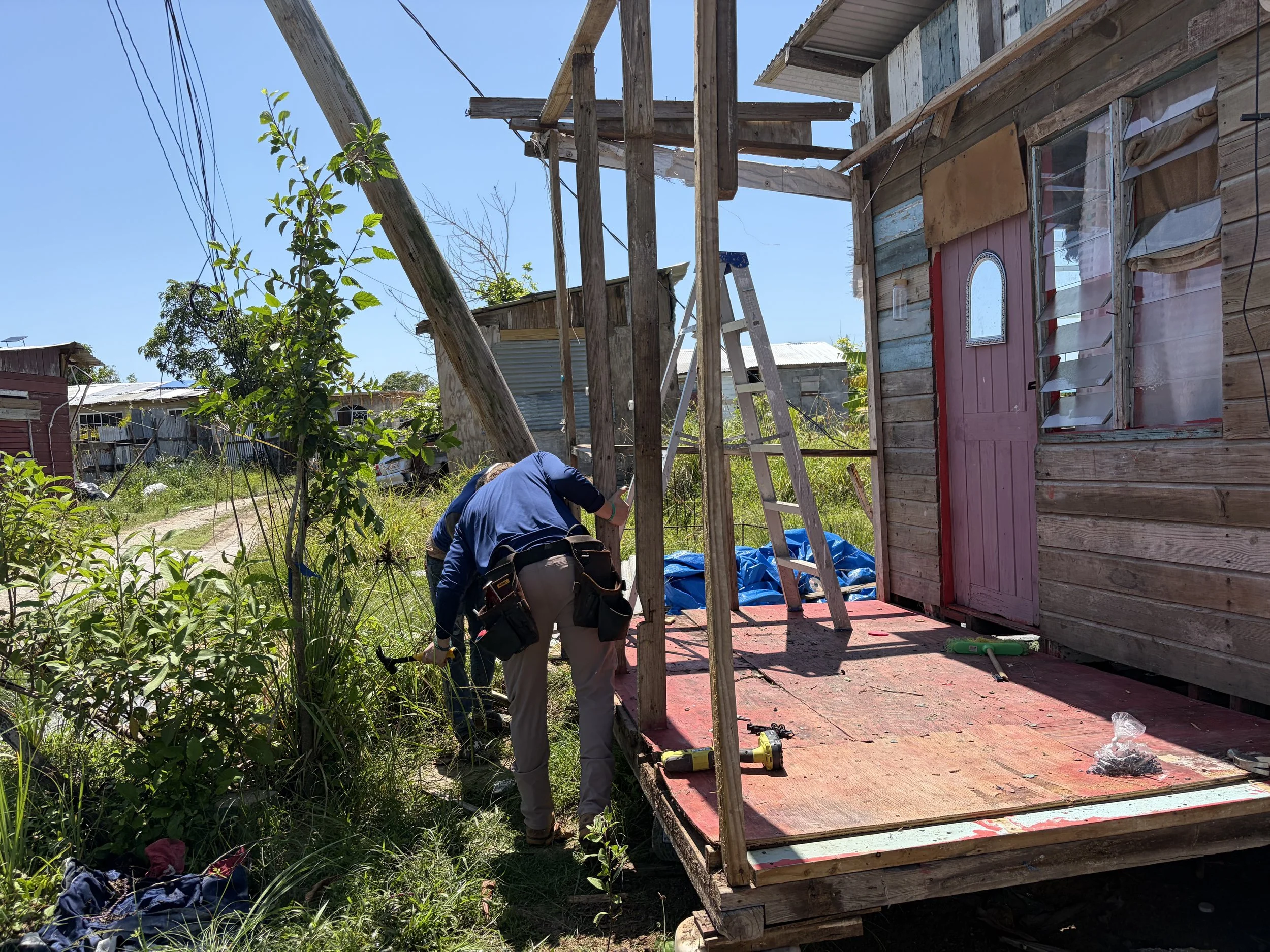 Person working on constructing a wooden house frame outdoors, with tools and building materials around, greenery and other houses in the background.