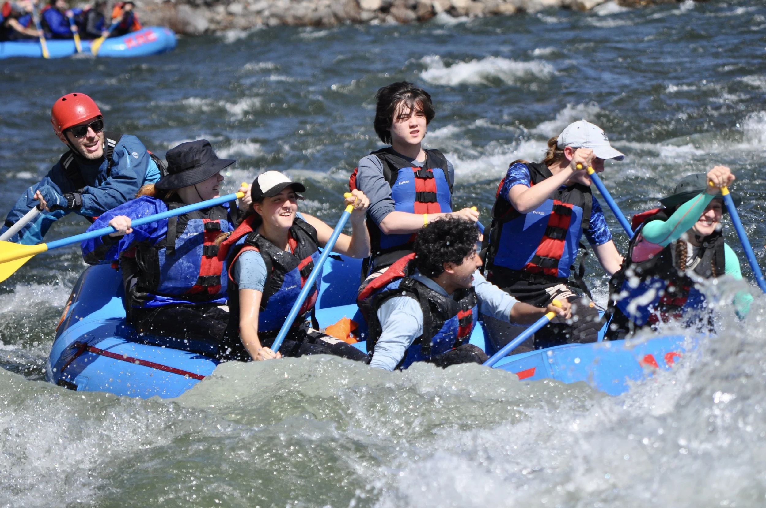 Group of people white water rafting on a river, wearing life jackets and holding paddles, with some wearing hats and helmets.