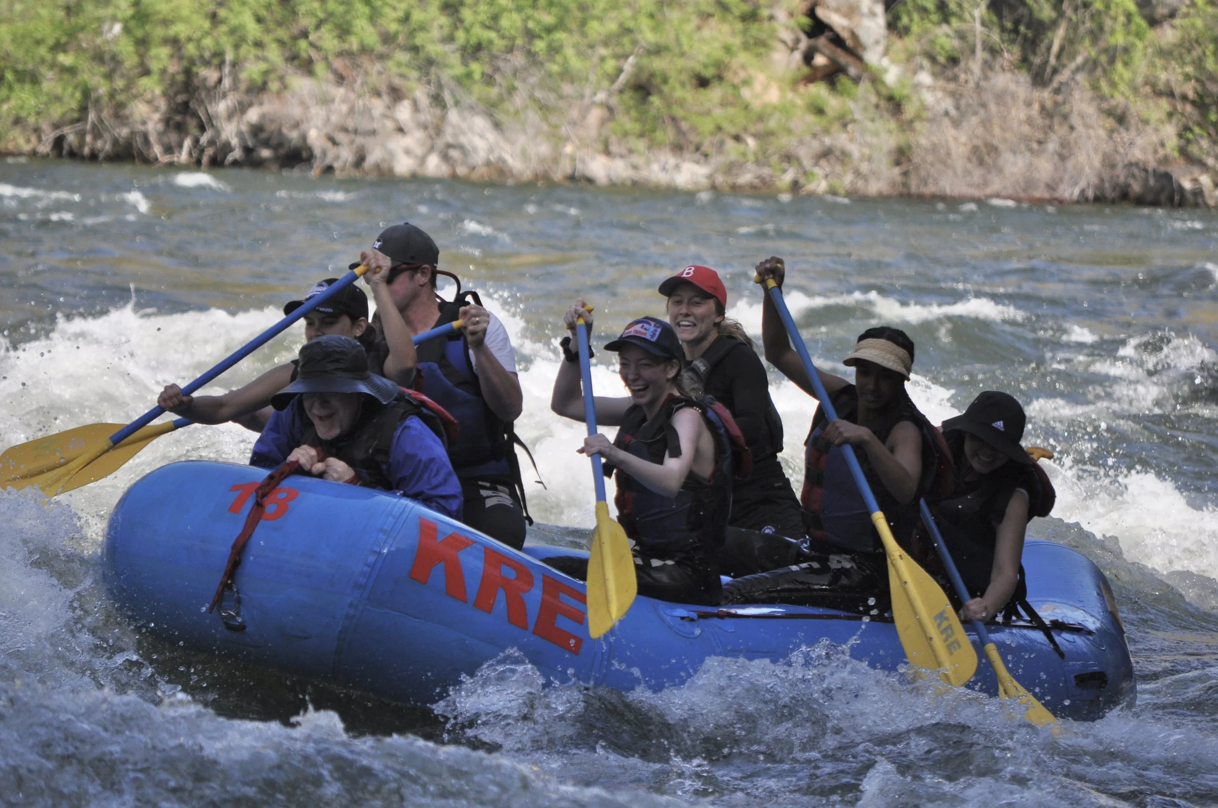 Group of people white water rafting on a river, smiling, holding paddles, wearing life jackets and helmets, with green trees in the background.