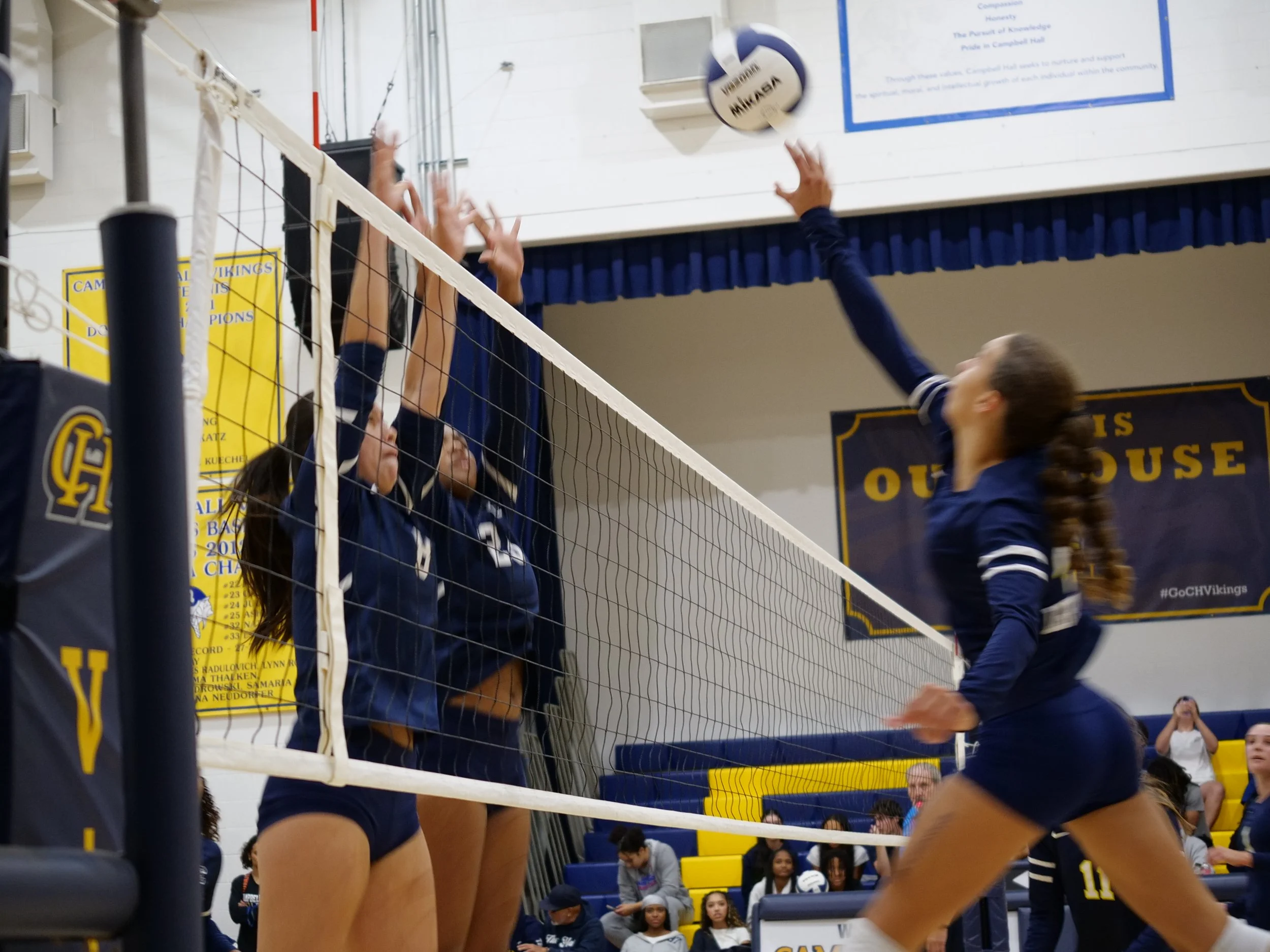Girls' volleyball game in an indoor gym, players jumping near the net, with spectators watching in the background.