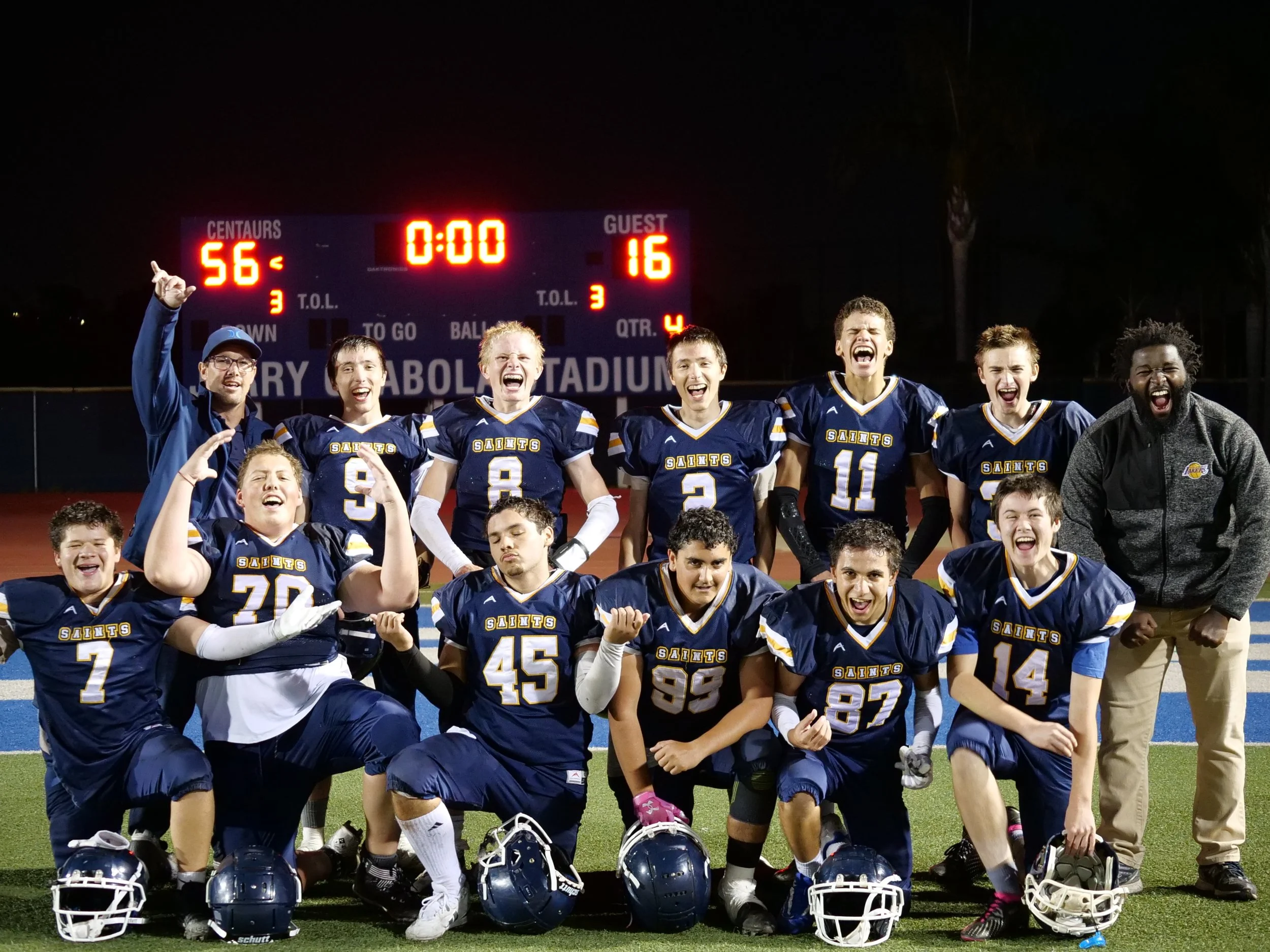 A group of football players and coaches celebrating on a football field at night after a game, with a scoreboard showing a final score of 56-16.