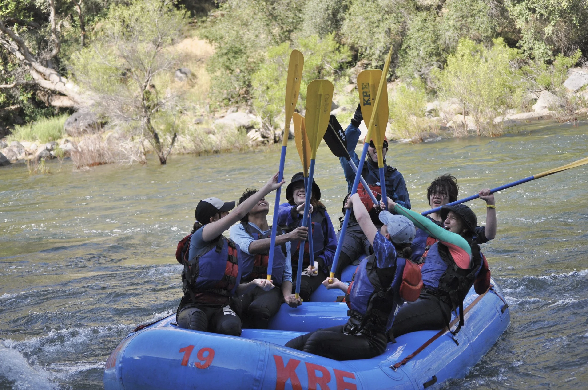 Group of people in a blue raft on a river, holding paddles and smiling, surrounded by green trees and rocks.