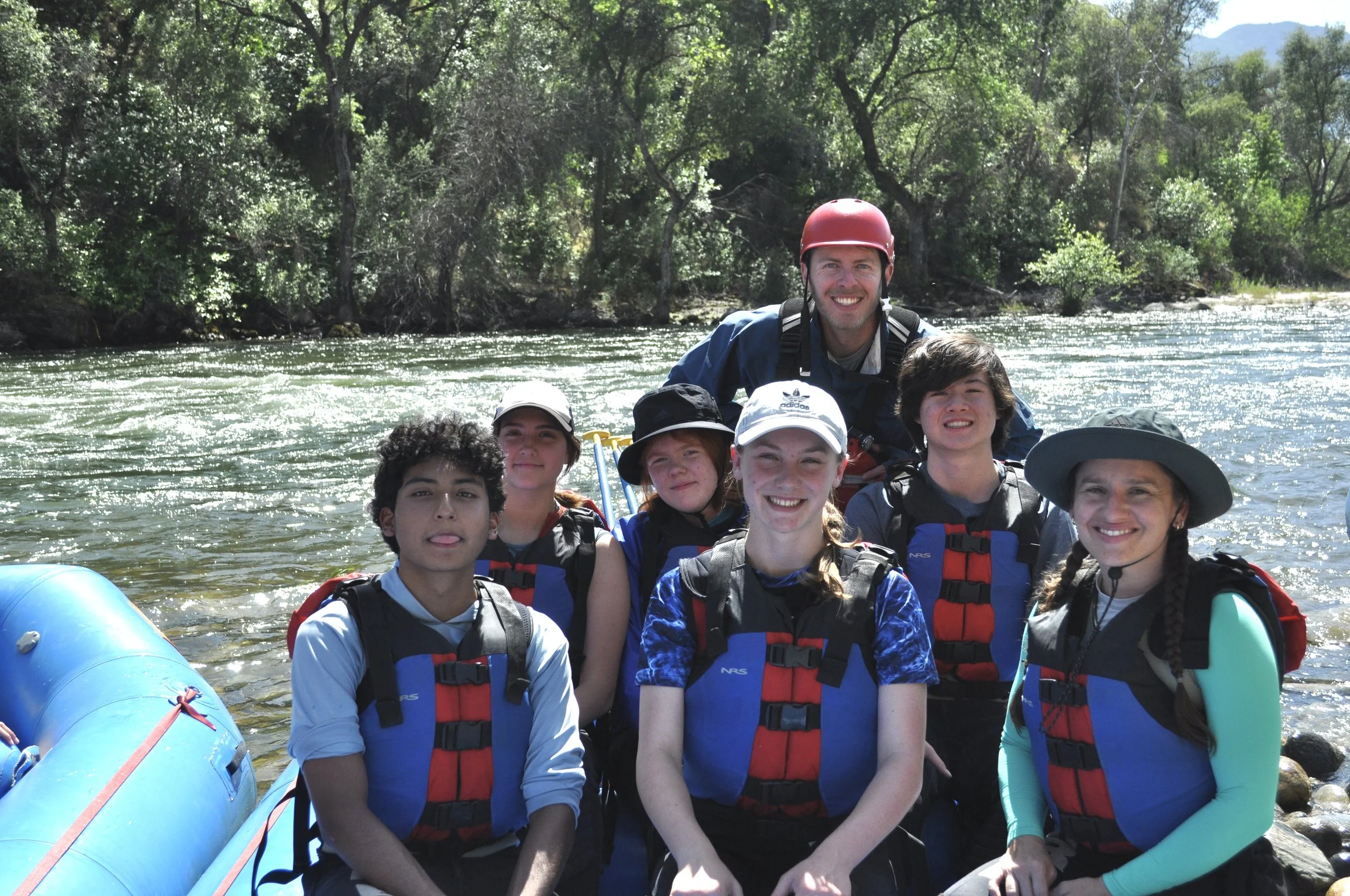Group of people on a boat in a river surrounded by lush green trees, with mountains in the background. They are wearing life jackets and smiling.