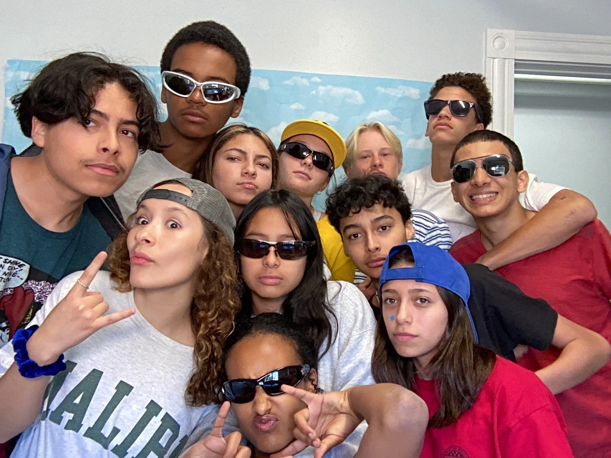 Group of teenagers wearing sunglasses, making gestures, and posing together indoors before a painted sky backdrop.