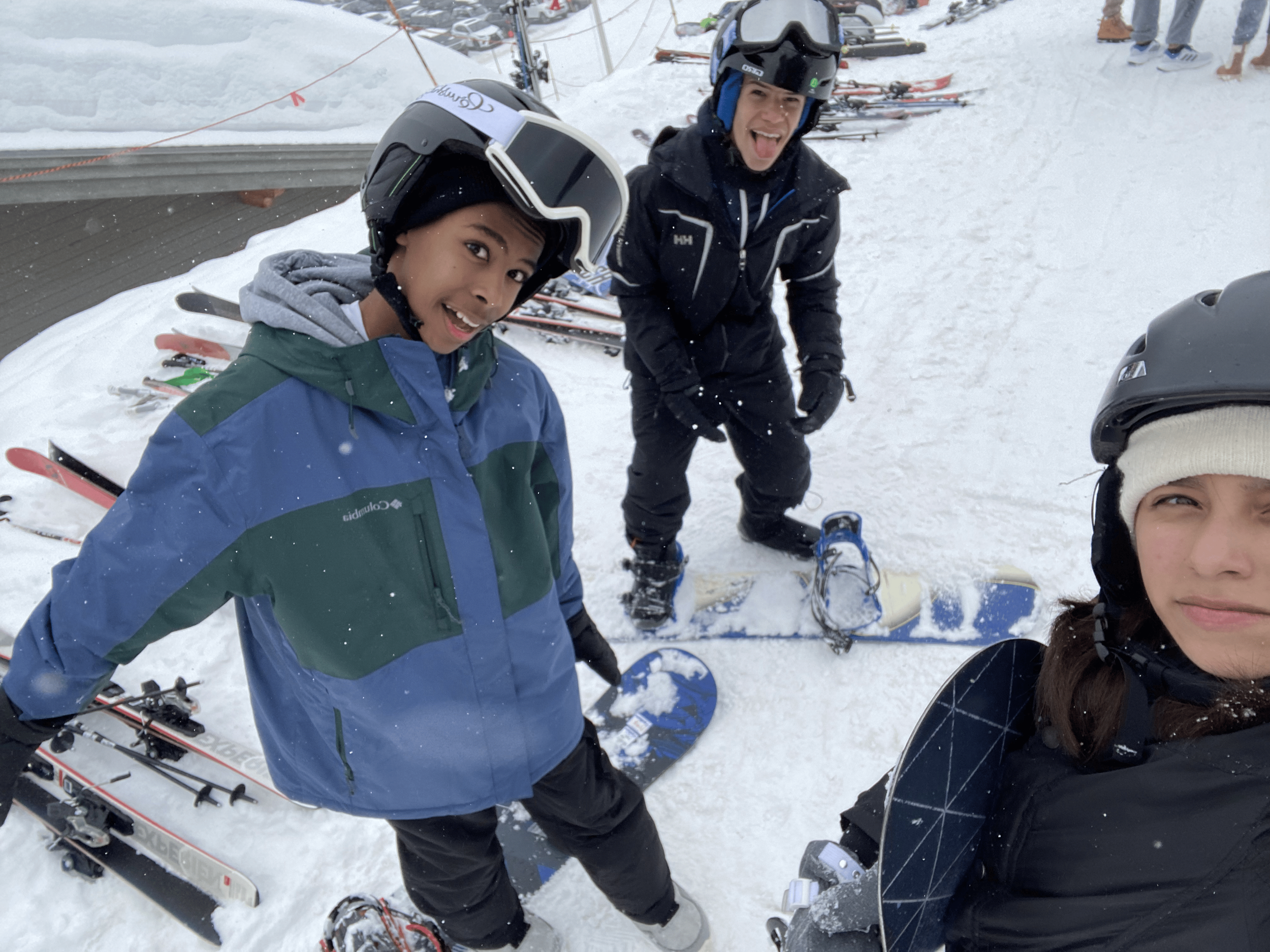 Two children smiling in ski gear on a snowy slope, with skis and helmets, and a group of people in the background