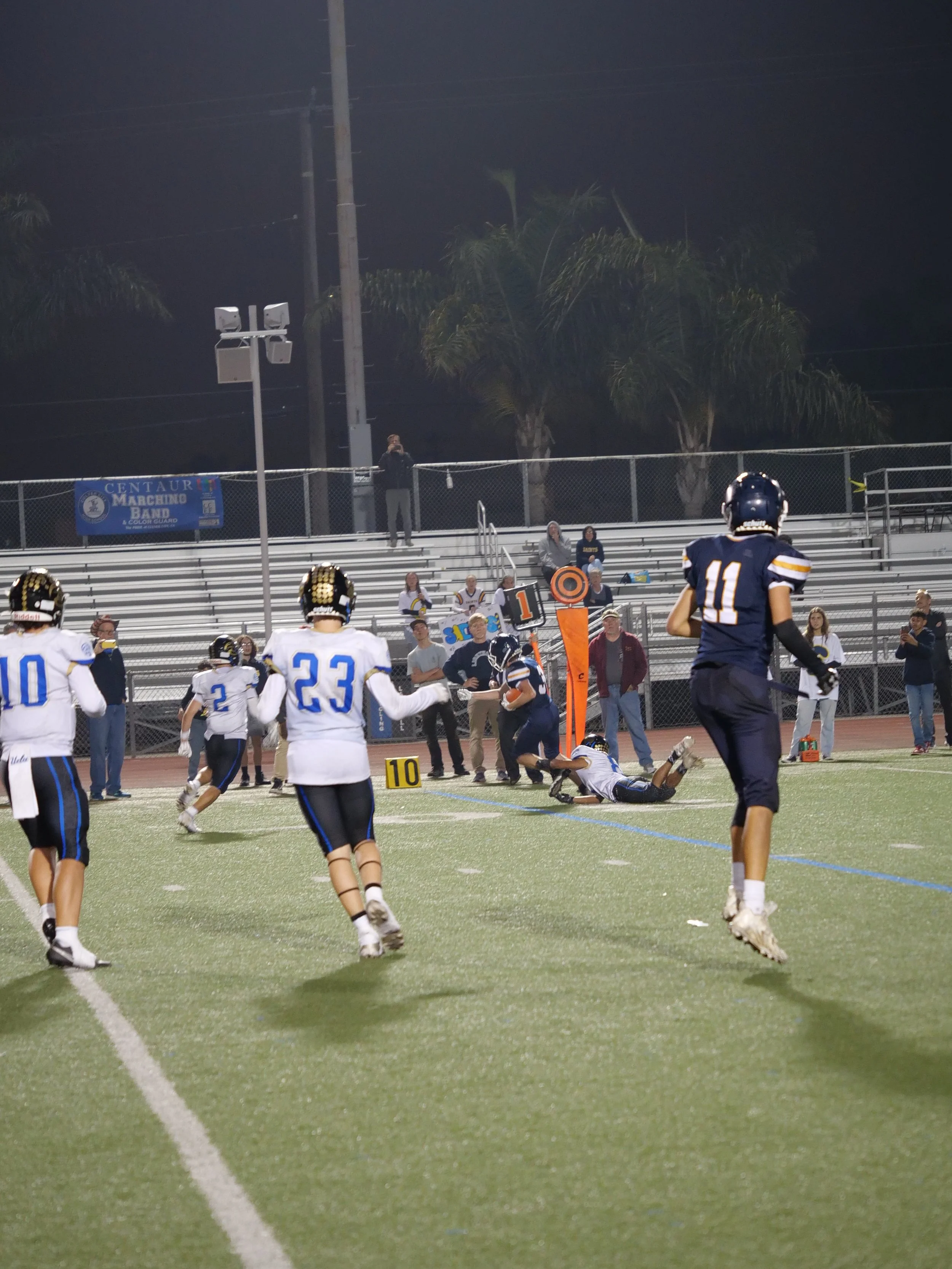 Nighttime football game with players and cheerleaders on a field, scoreboard, and spectators on the bleachers.