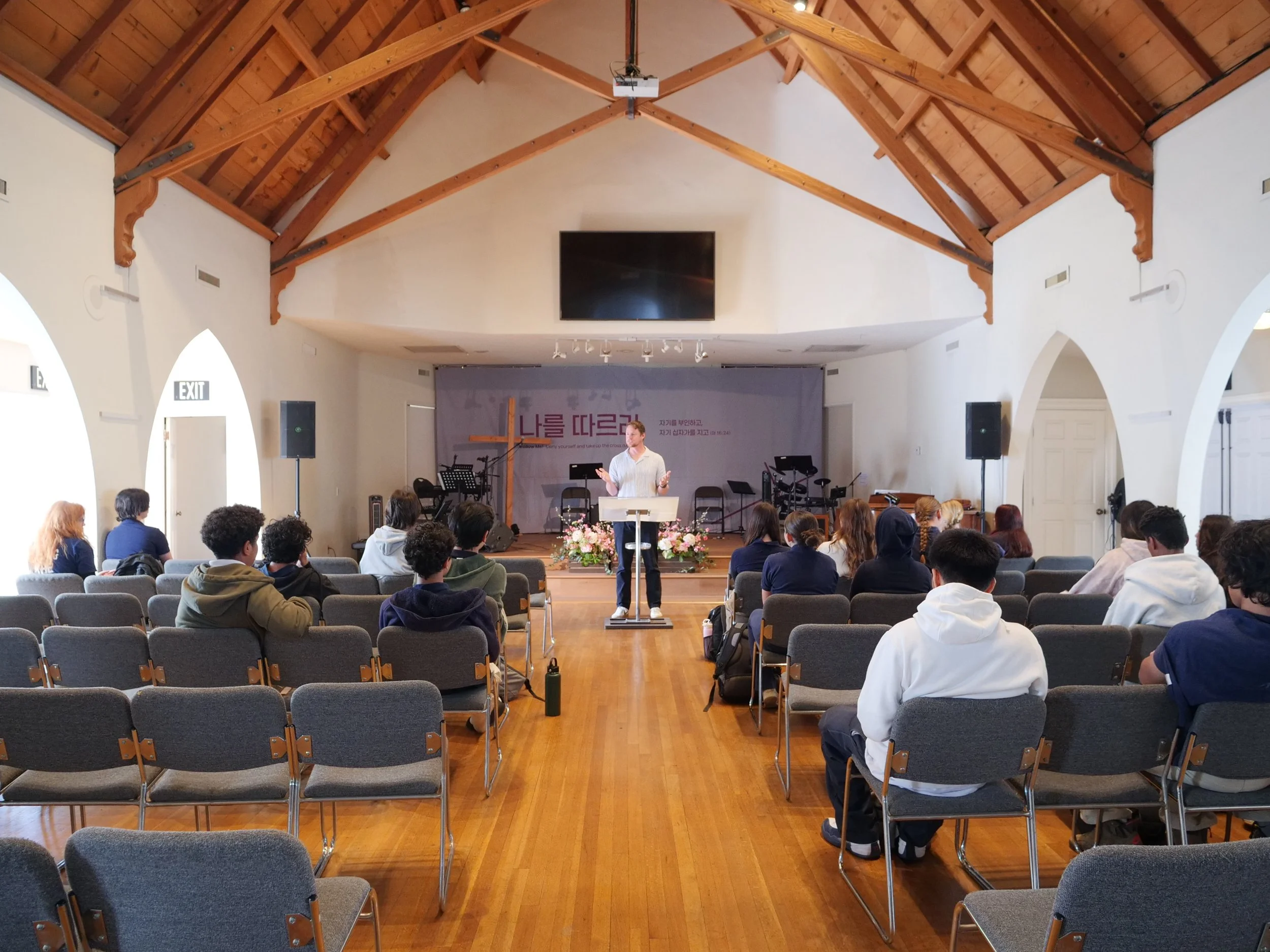 A man standing behind a podium giving a talk in a church or conference room with wooden ceiling beams and a large screen on the wall behind him. Attendees are sitting in rows of chairs facing the front.