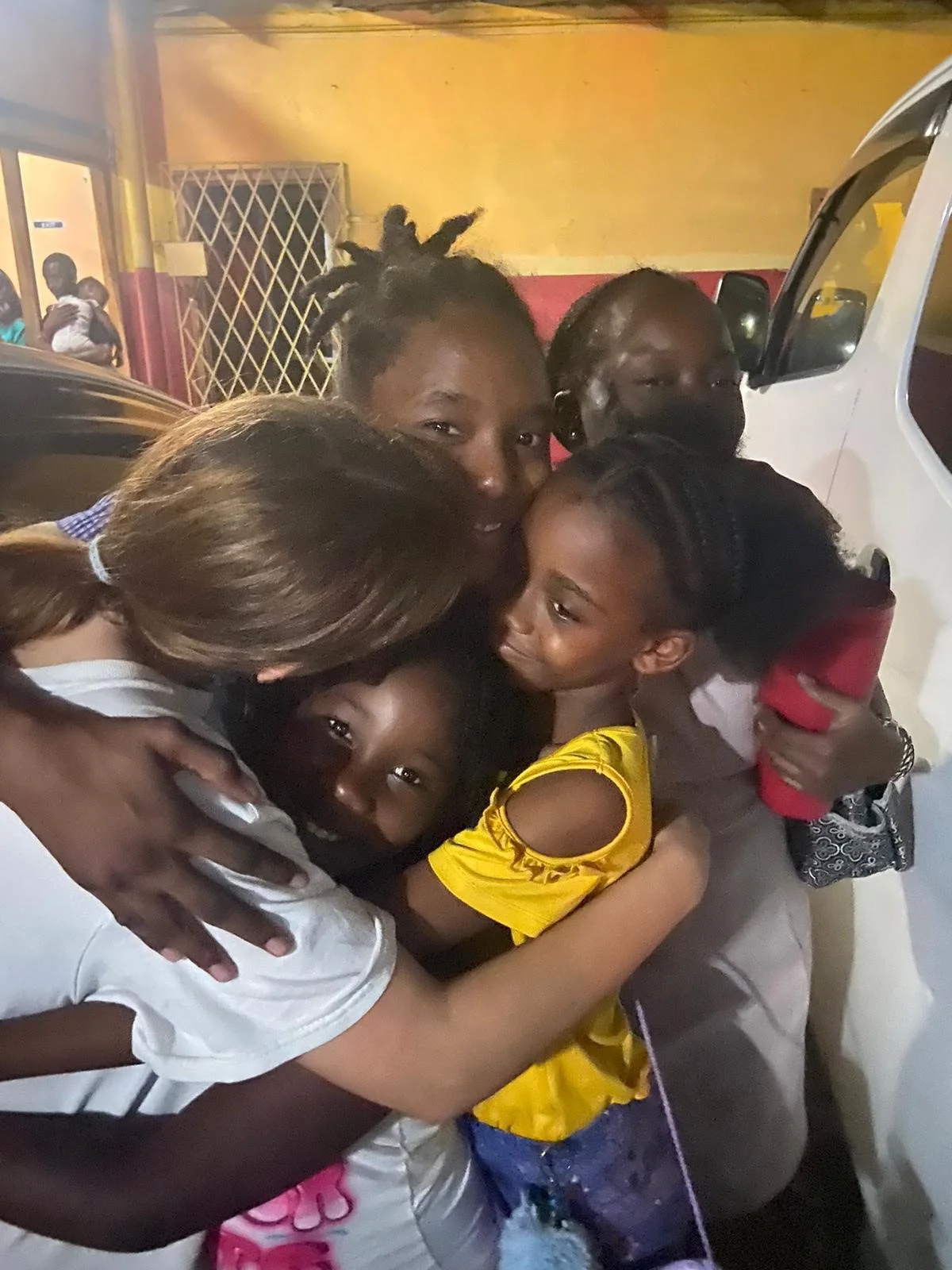Group hug of smiling children and women outdoors at night, with a yellow and red wall and a white vehicle in the background.