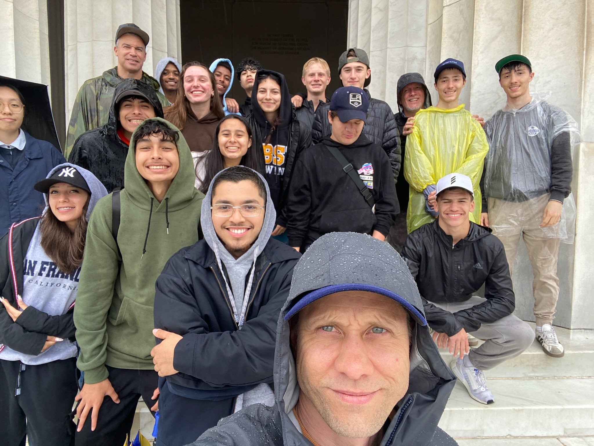 A group of diverse people, young and old, gathered on steps in front of a building, smiling for a selfie, some wearing rain jackets or hoodies, with overcast weather and raindrops on the camera lens.