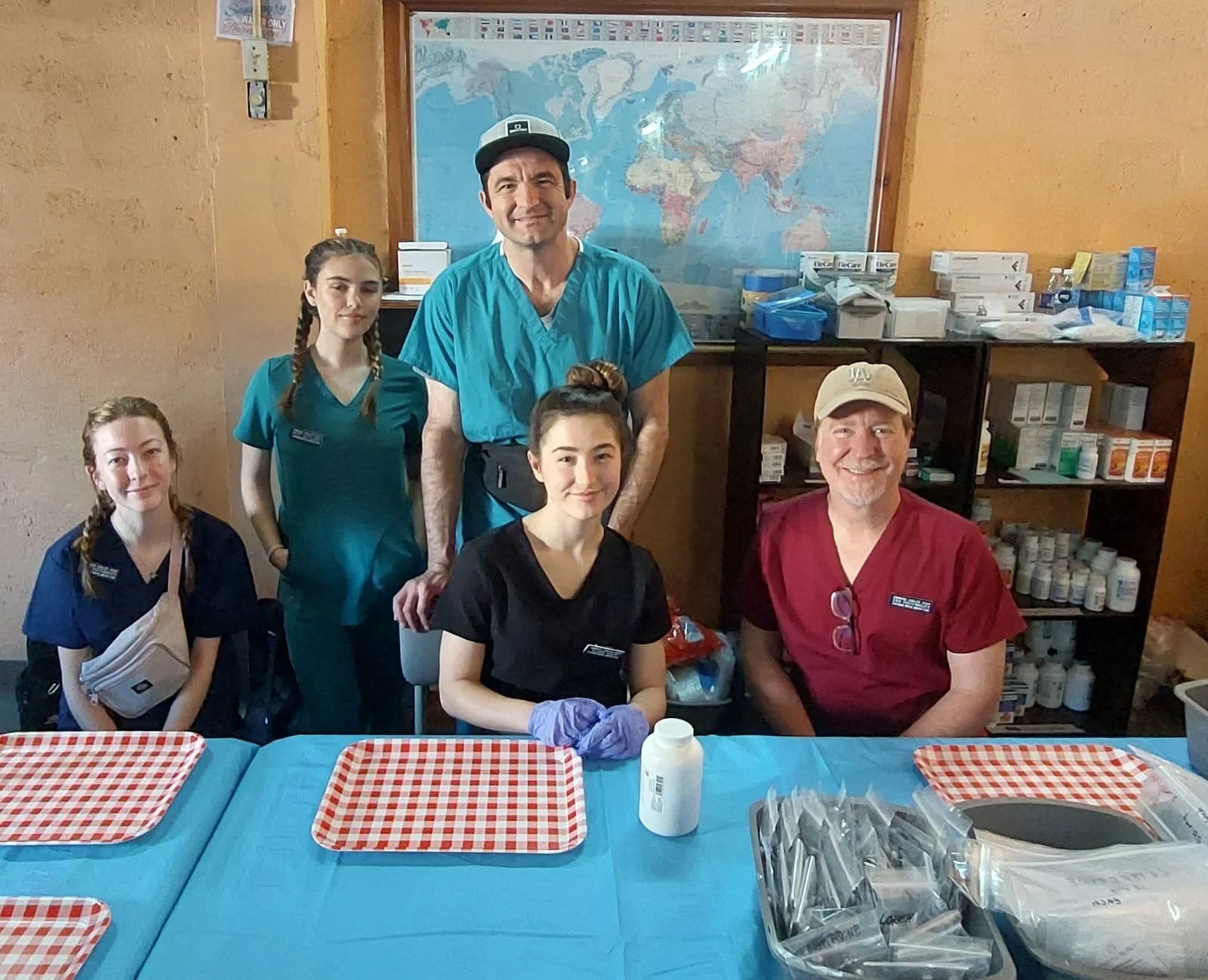 Group of five healthcare workers in scrubs and uniforms in a room with a world map on the wall and shelves of medical supplies, standing and sitting behind a table with checked paper trays and medical equipment.