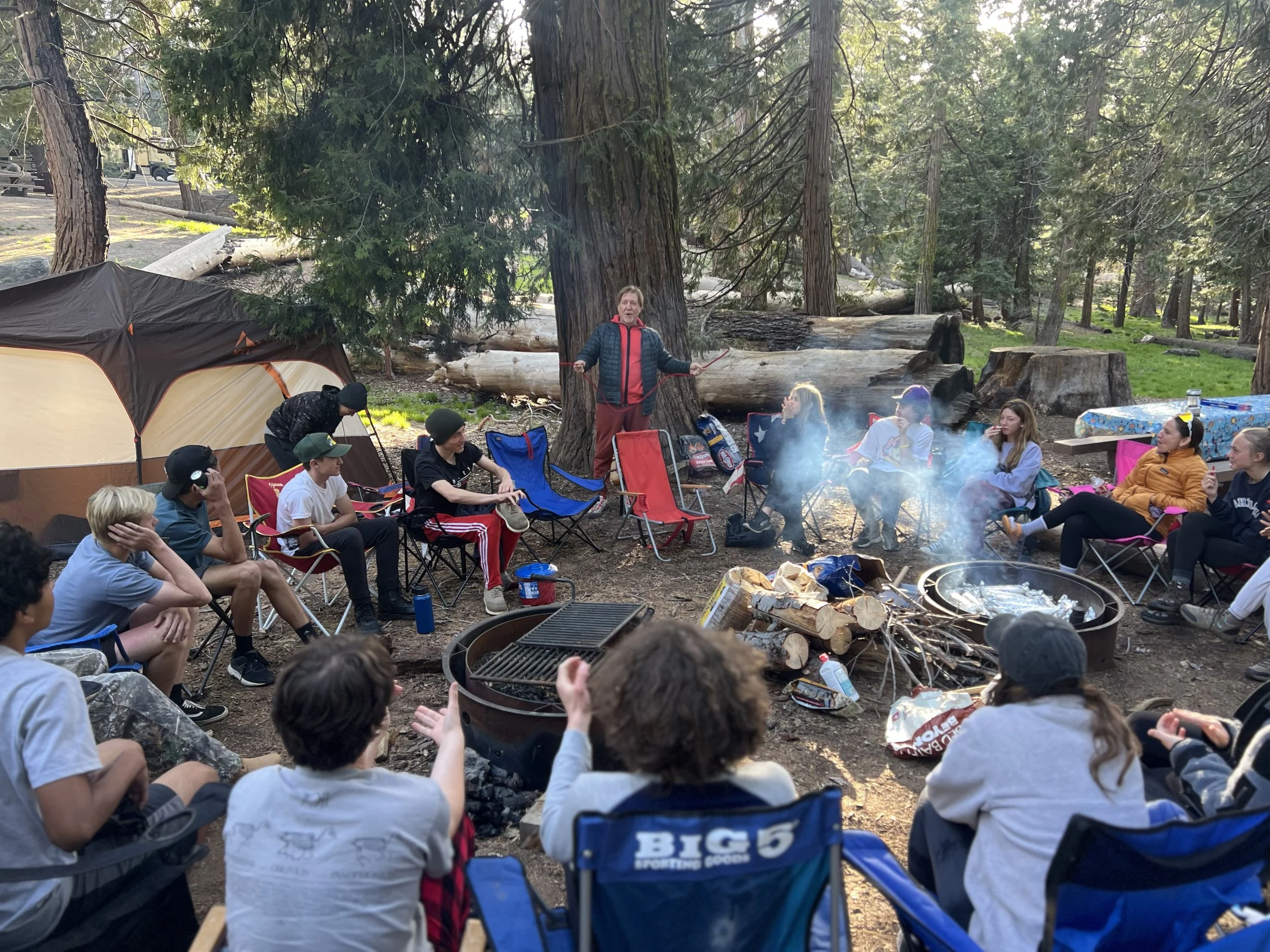Group of people camping in a forest, sitting around a campfire with a woman standing and speaking, tents, and fallen logs visible in the background.