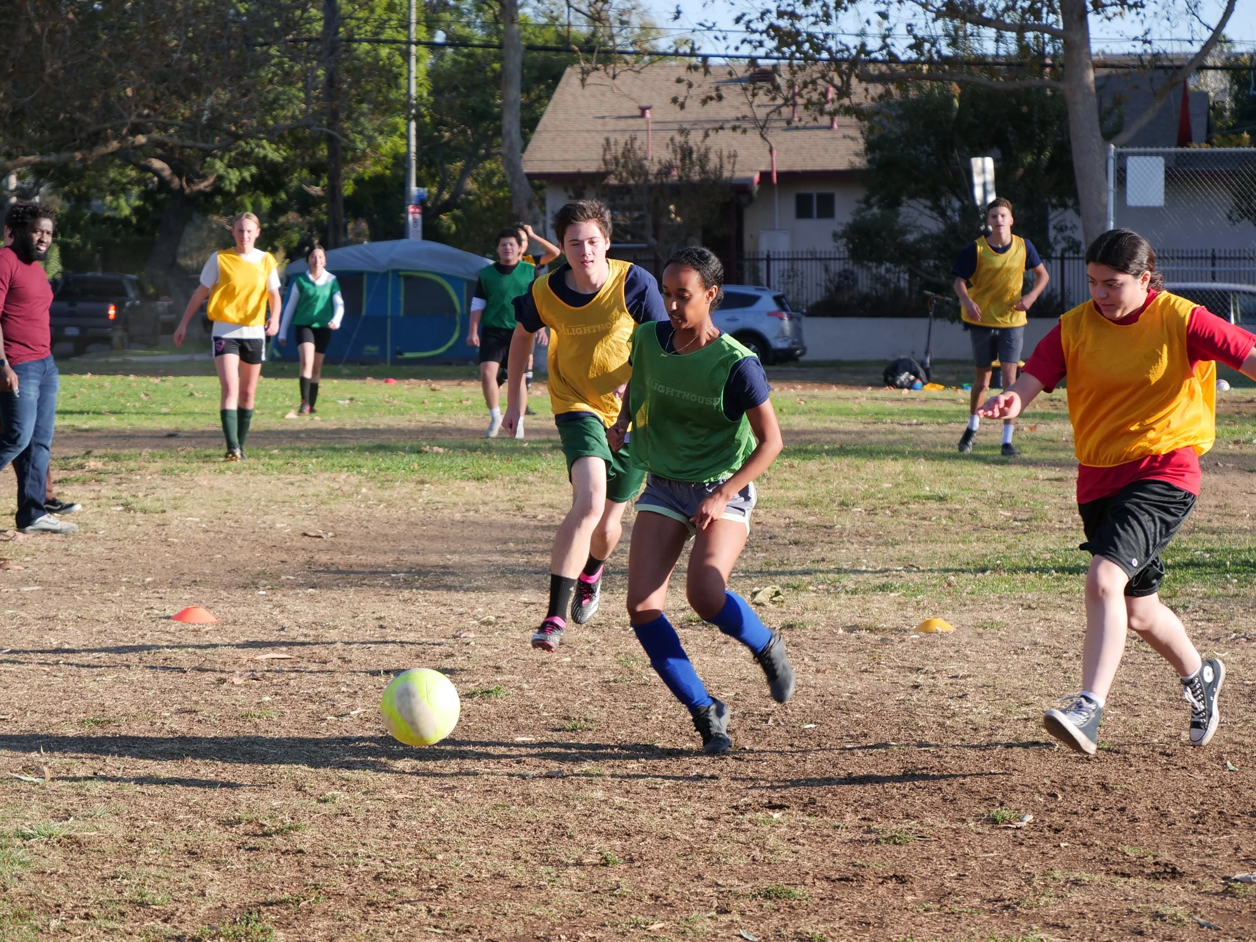 A group of children playing soccer outdoors on a grassy field, with some wearing yellow and others green jerseys. They are running towards a white and yellow soccer ball, with residential houses and trees in the background.