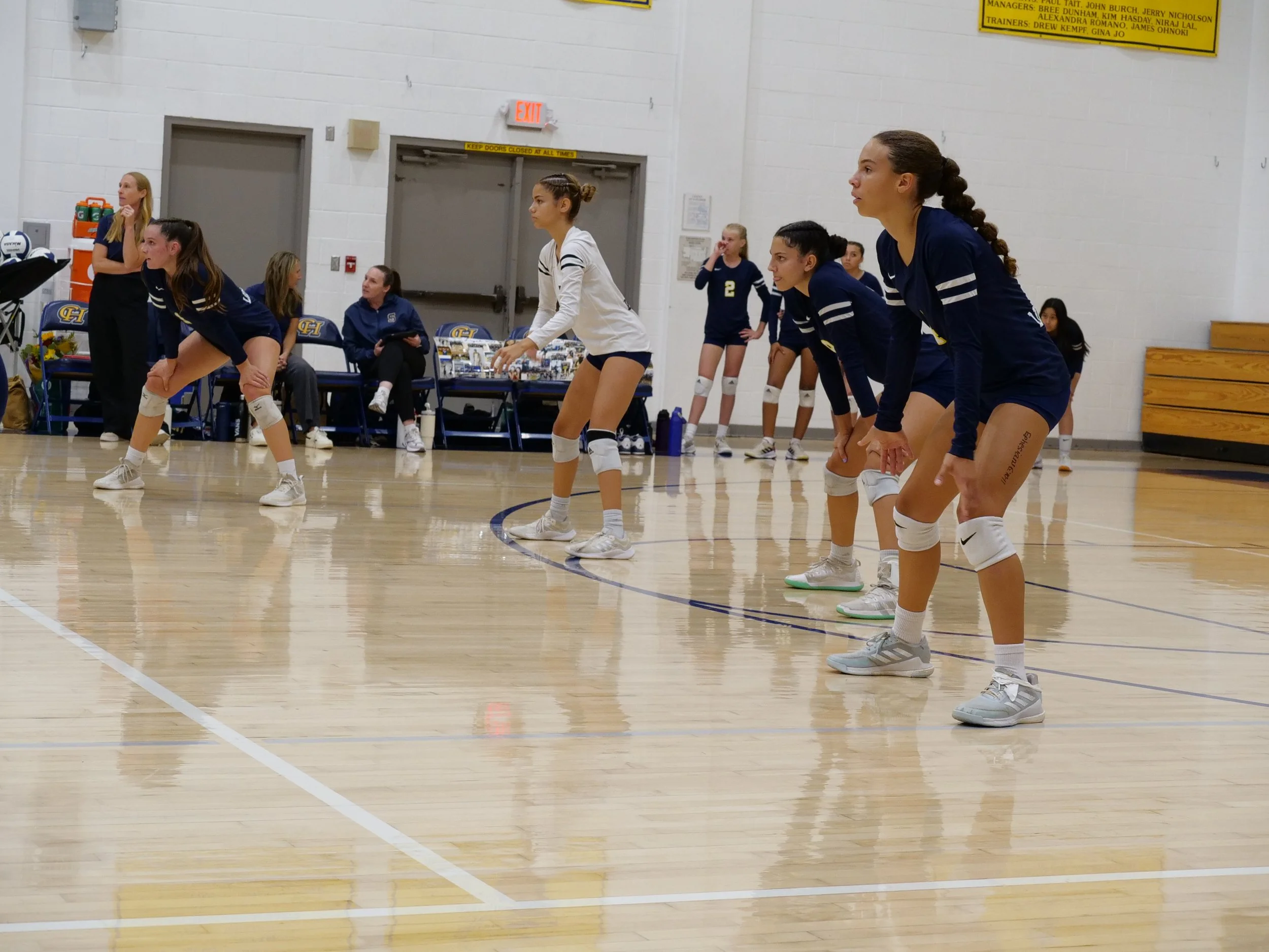 Women volleyball players in uniform standing in a gymnasium during a team huddle or pre-game warm-up.