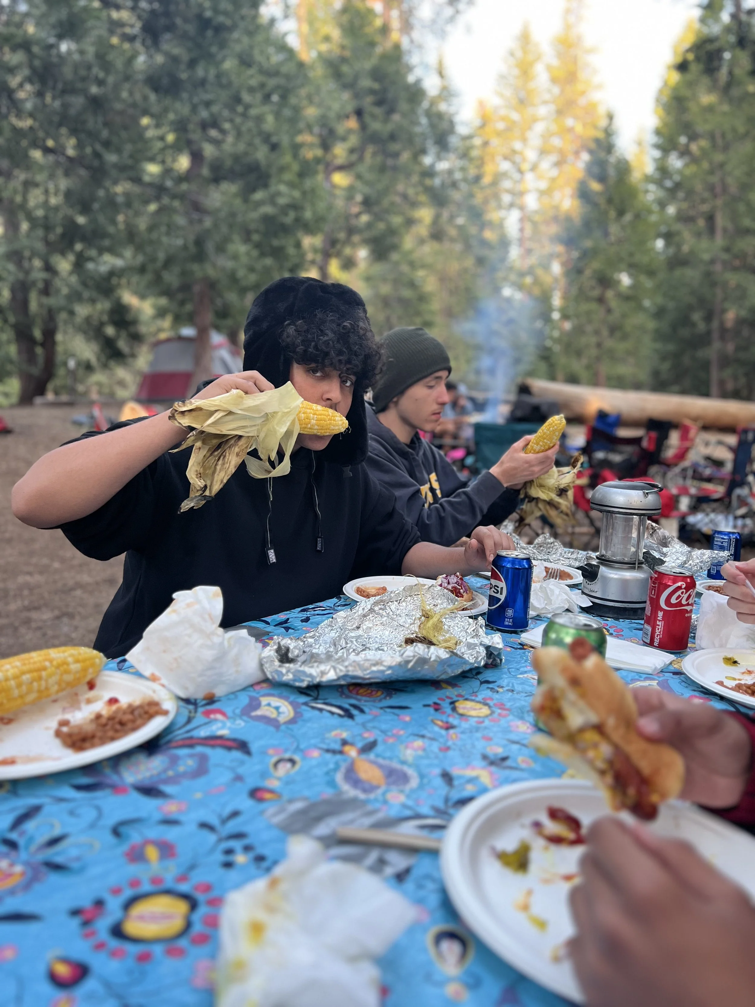 People sitting at an outdoor table with food, including corn on the cob, nachos, and drinks, in a wooded area.