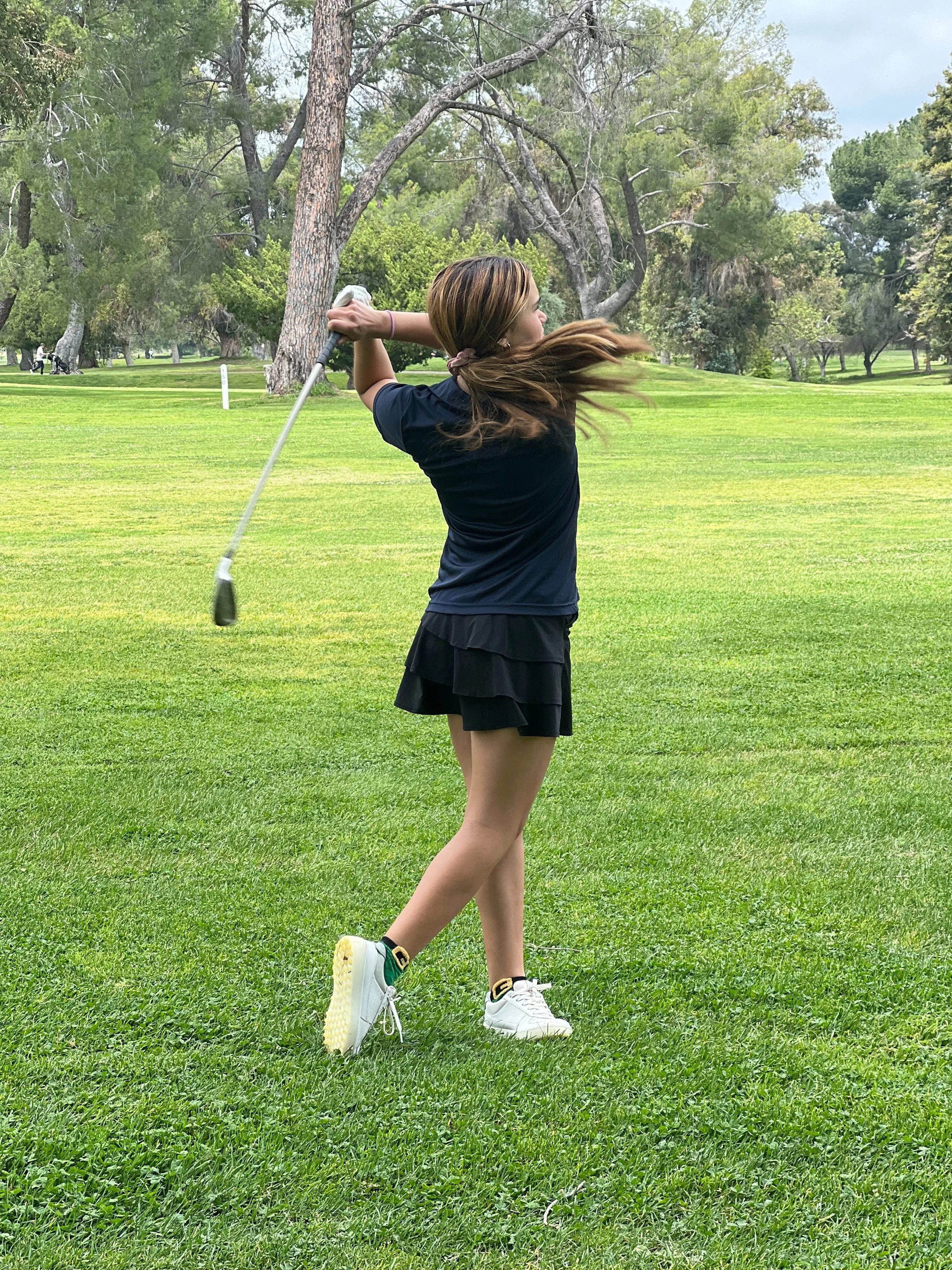 A woman playing golf on a lush green golf course with trees in the background, captured from the side as she swings a golf club.