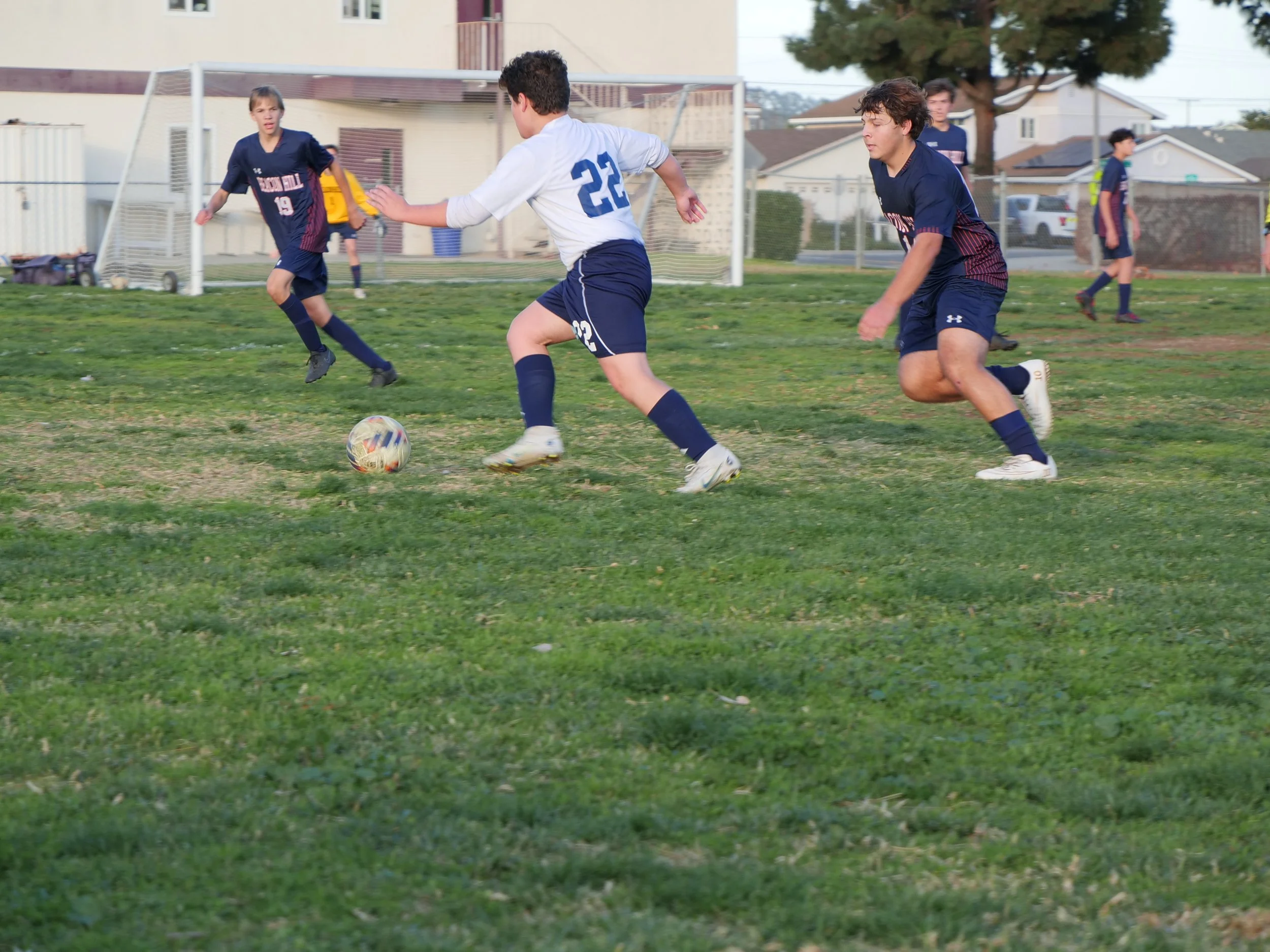 Young boys playing soccer on a grassy field, some running after the ball, with a soccer goal and houses in the background.