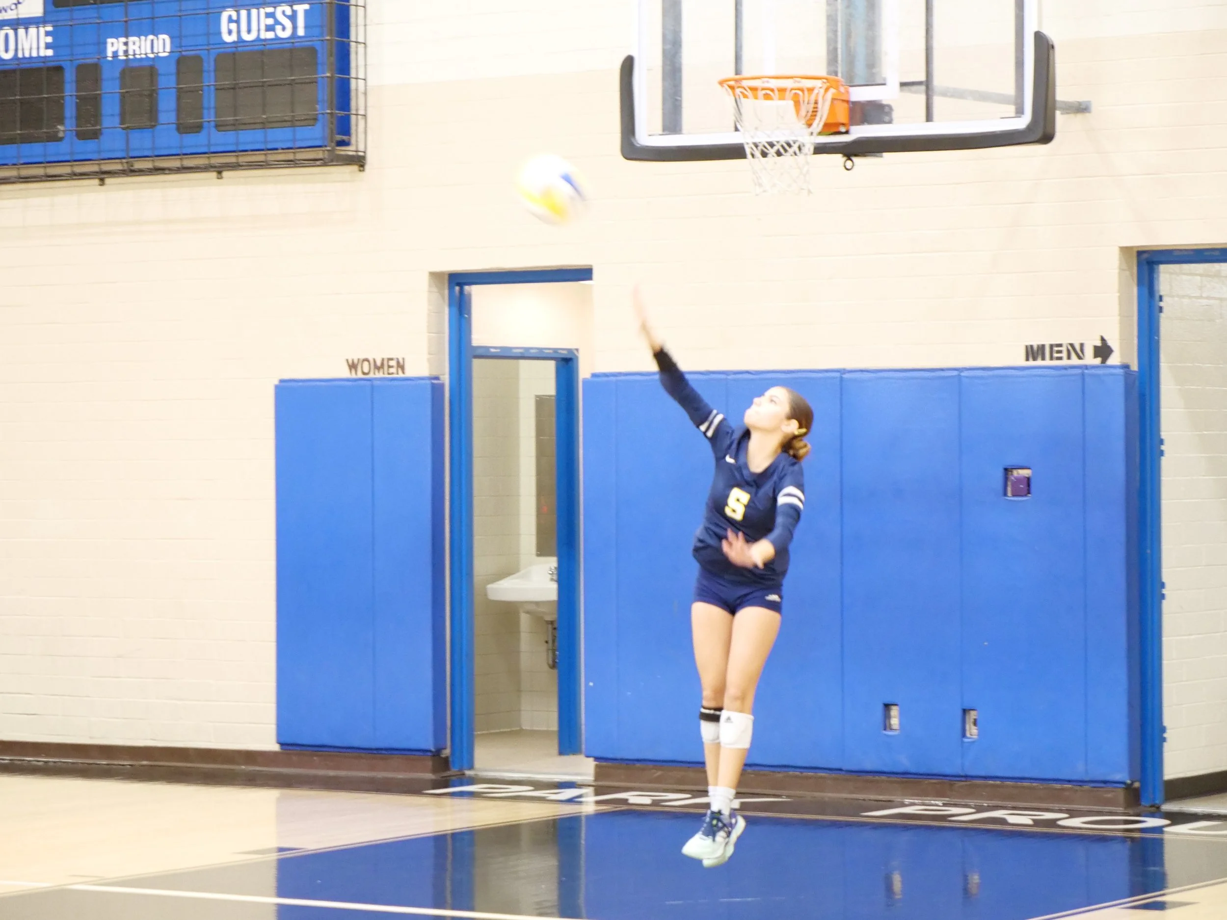 A girl playing volleyball inside a gymnasium, jumping to hit the ball toward the basketball hoop. Background includes a women's restroom sign and a men’s restroom sign on the wall.