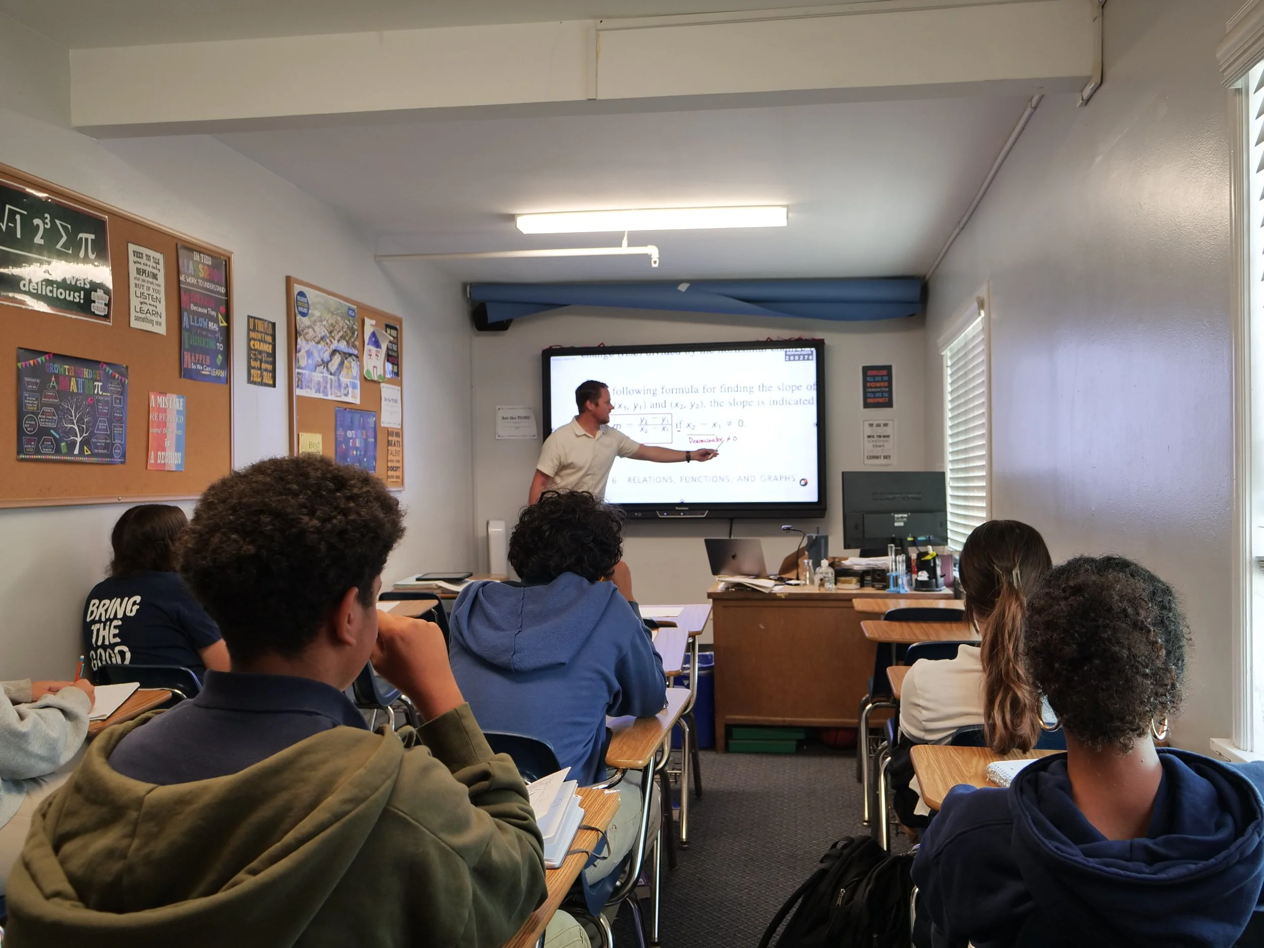 A classroom with students seated at desks, listening to a teacher pointing at a large screen displaying mathematical formulas. Posters and academic materials are on the walls, and sunlight is coming through window blinds.