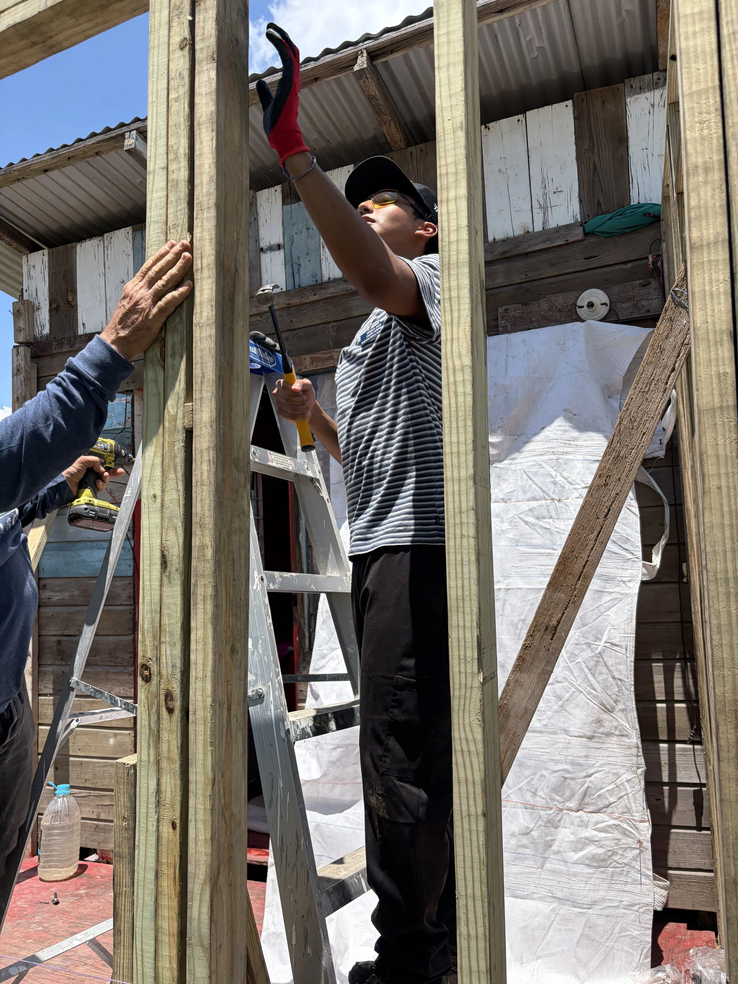 Man working on wooden framing for a building under construction, wearing sunglasses, a striped shirt, and a cap, with a ladder and construction tools nearby.