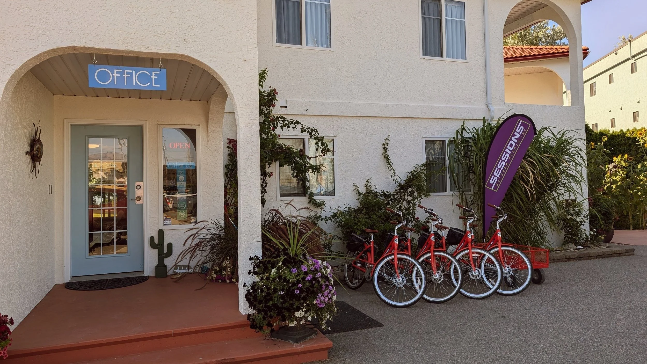 A white building with an arched entrance labeled 'OFFICE' with a blue sign. There are four red bikes with white wheels and a purple sign that says 'SESSIONS' outside, surrounded by plants and flowers.