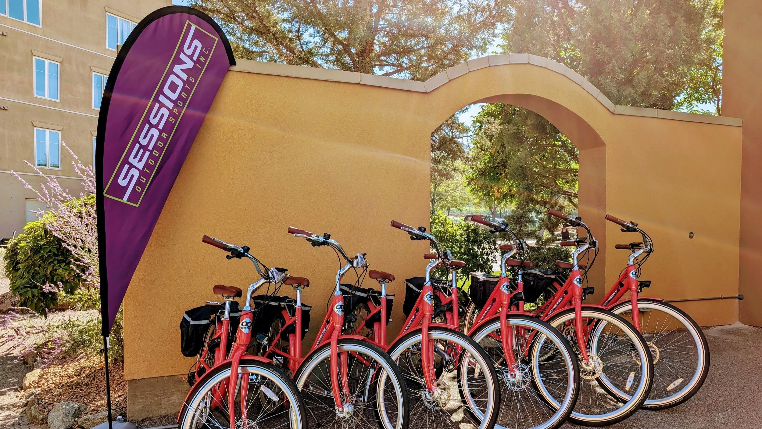 Six red rental bicycles parked side by side outdoors beside a yellow stucco wall with an archway and a purple banner that reads 'SESSIONS' in white letters, with trees and a building in the background.