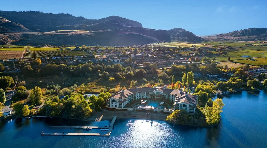 Aerial view of a lakeside residential community surrounded by trees, with a body of water in the foreground and rolling hills and farmland in the background.