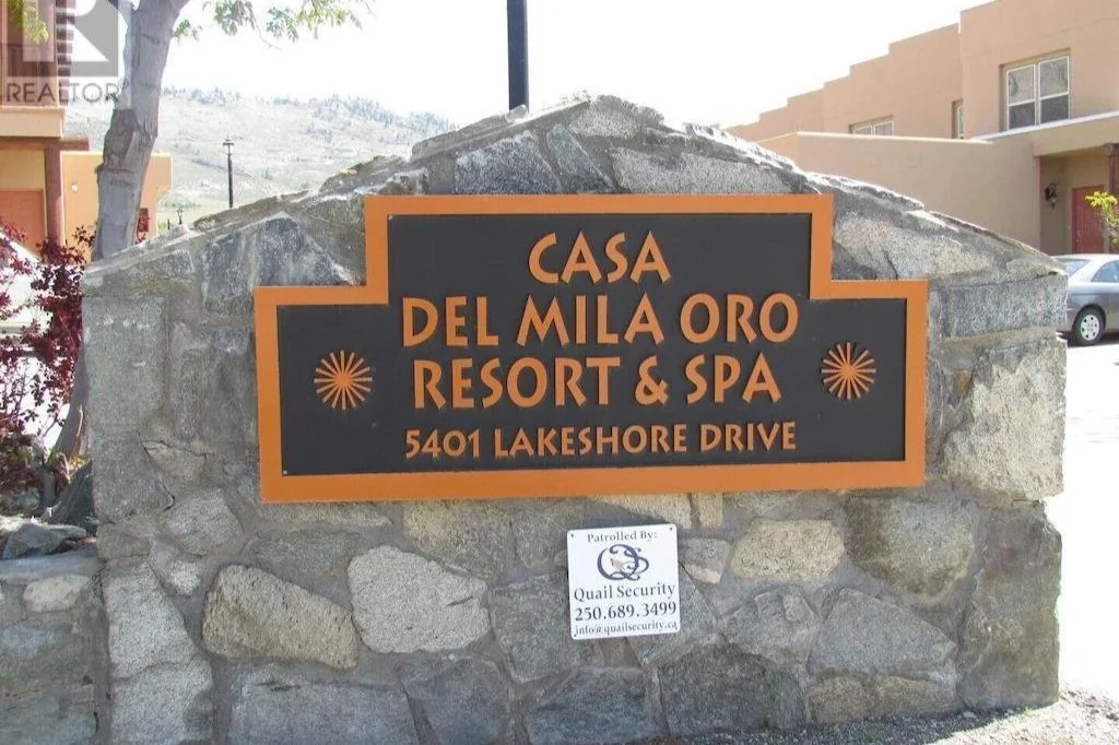 A stone sign with an orange border and black background displaying the text, 'Casa Del Mila Oro Resort & Spa, 5401 Lakeshore Drive.' Below the sign, there is a smaller white sign indicating security patrol.