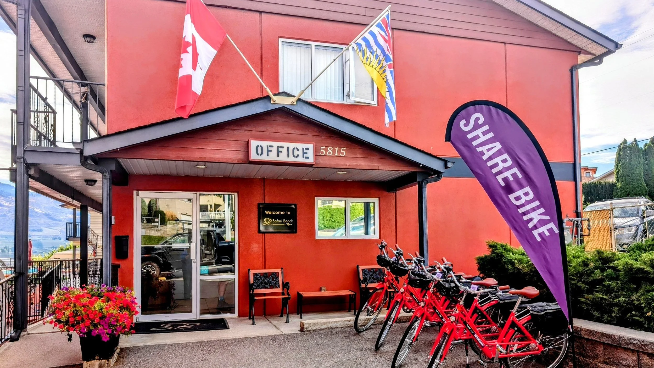 The entrance of a red building with the word 'OFFICE' and the number 5815. There is a purple flag with 'SHARE BIKE' written on it, several red bicycles parked outside, two chairs, a potted pink flower, and Canadian and provincial flags flying above.