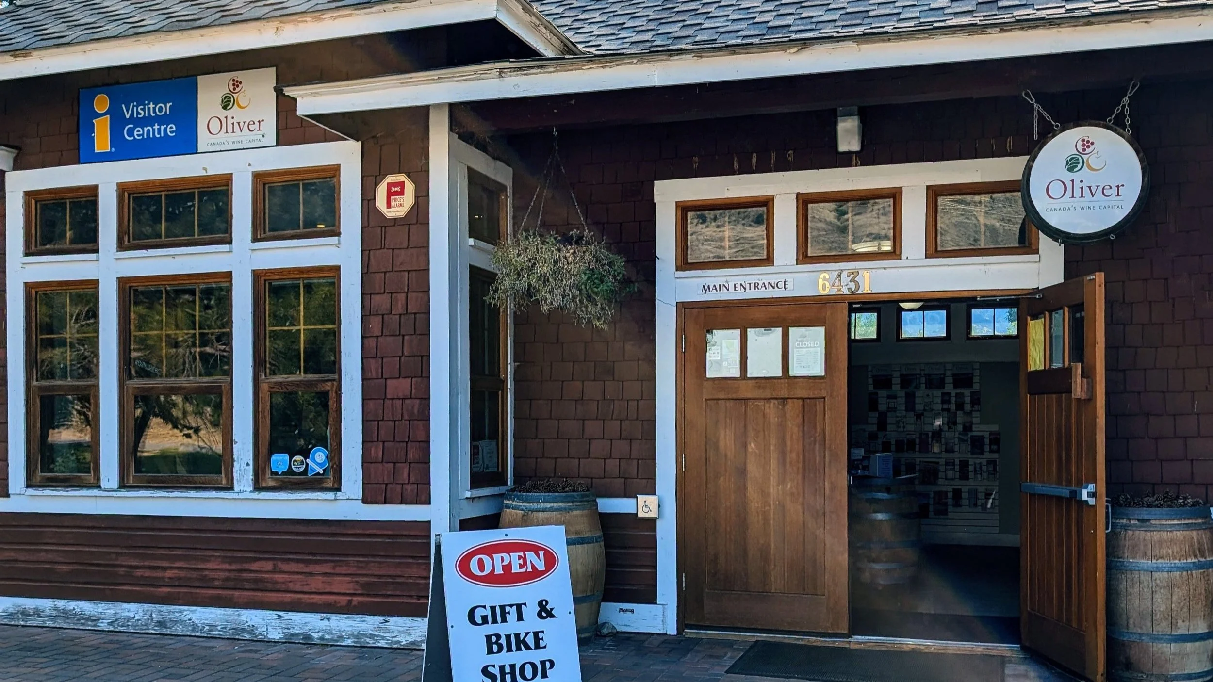 Exterior of a gift and bike shop with wooden door open, sign reading 'Open Gift & Bike Shop' outside, located at 6431 Oliver, Canada&aposs wine capital, with a sign indicating visitor centre and a hanging round sign for Oliver.