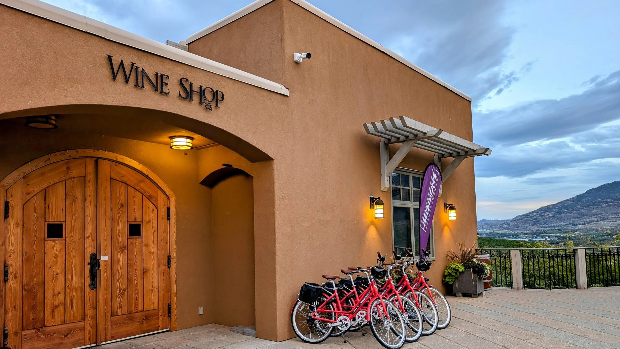 Exterior view of a wine shop with a large wooden door, warm lighting, and four red bicycles parked outside. There are potted plants, a purple flag, and scenic mountains in the background.