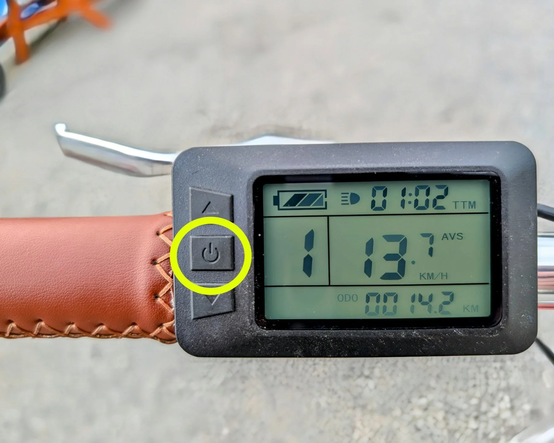 Digital bicycle speedometer mounted on a brown leather handlebar grip, displaying 13.7 km/h, 1 hour and 2 minutes, and 14.2 kilometers traveled. The power button is circled in yellow.