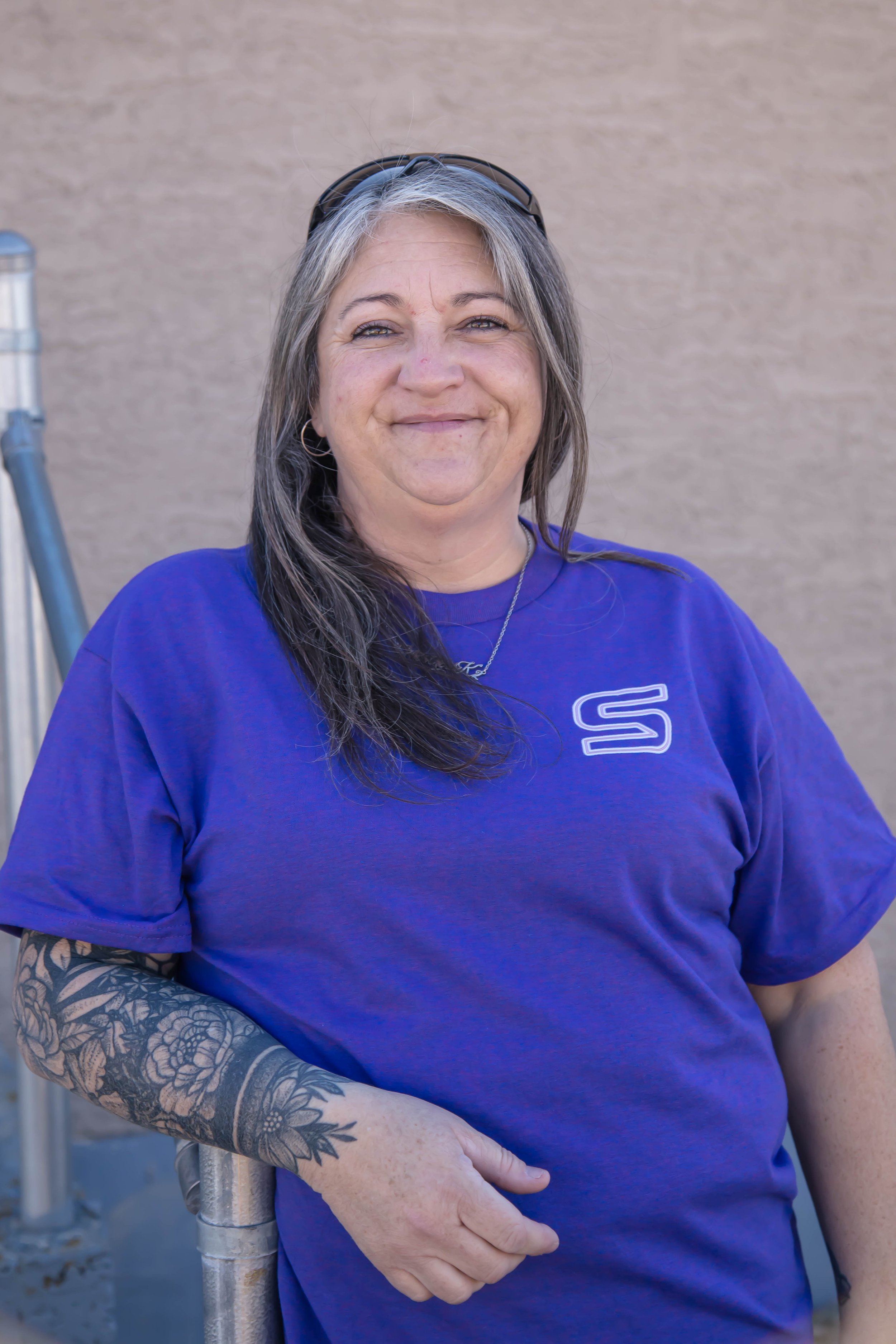 A smiling woman wearing a purple t-shirt with a logo, standing outdoors against a beige wall, with her left hand resting on a metal railing.