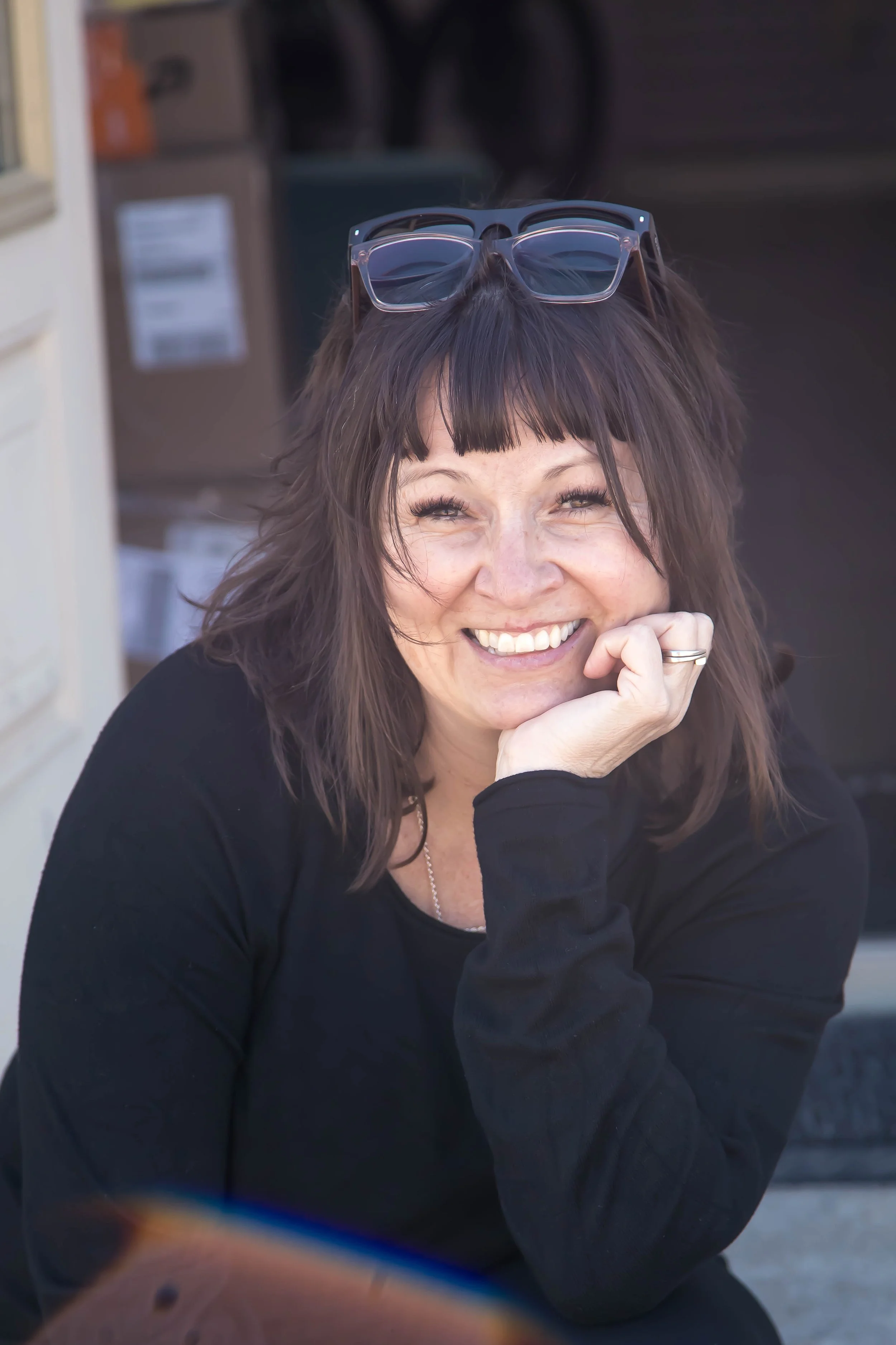 A woman with shoulder-length brown hair and bangs, wearing a black top, smiling with her hand resting on her chin, sunglasses on her head, outside in front of a doorway and cardboard boxes.