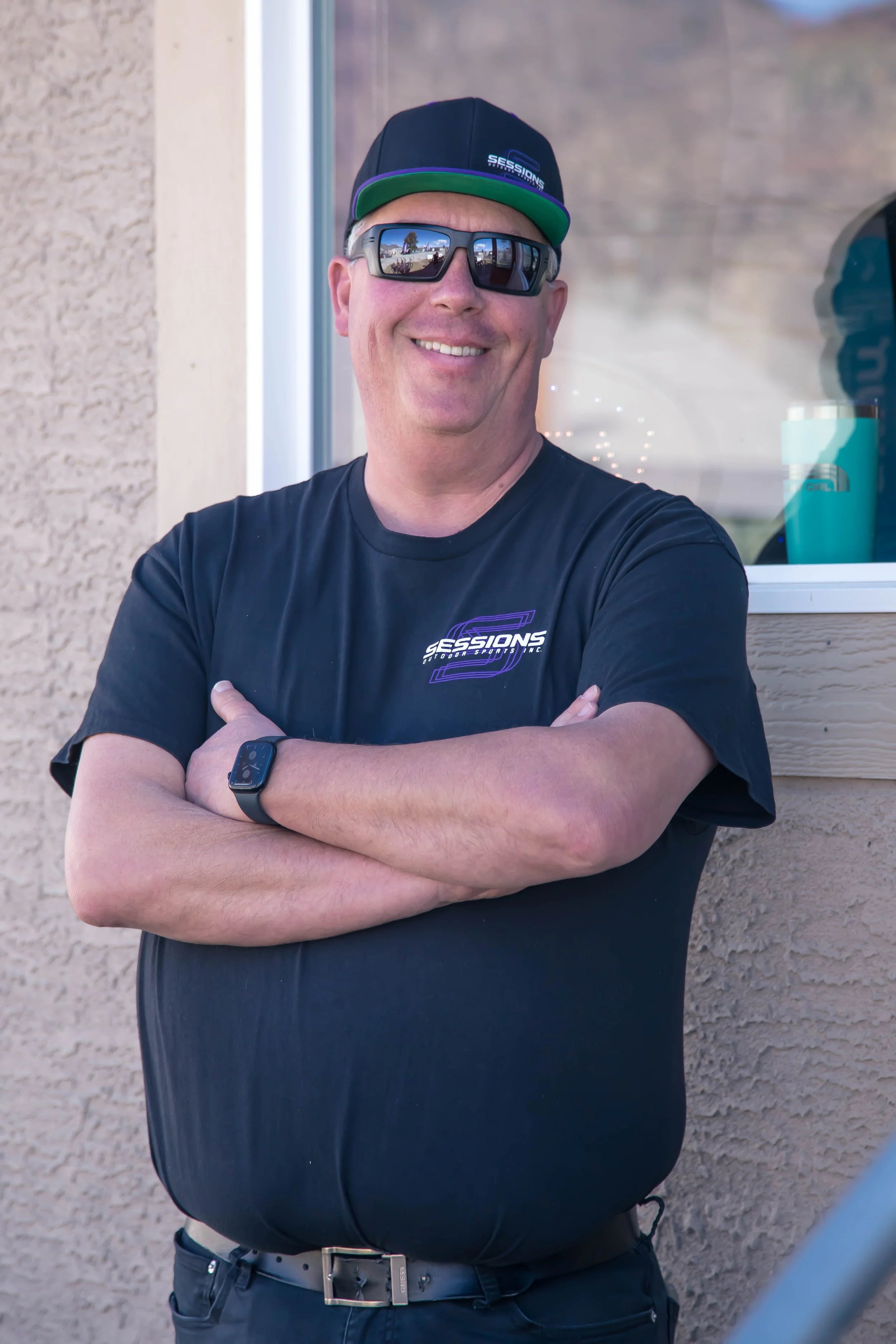 Smiling man wearing sunglasses, a black T-shirt, a cap, and a smartwatch, standing outside near a window with his arms crossed.