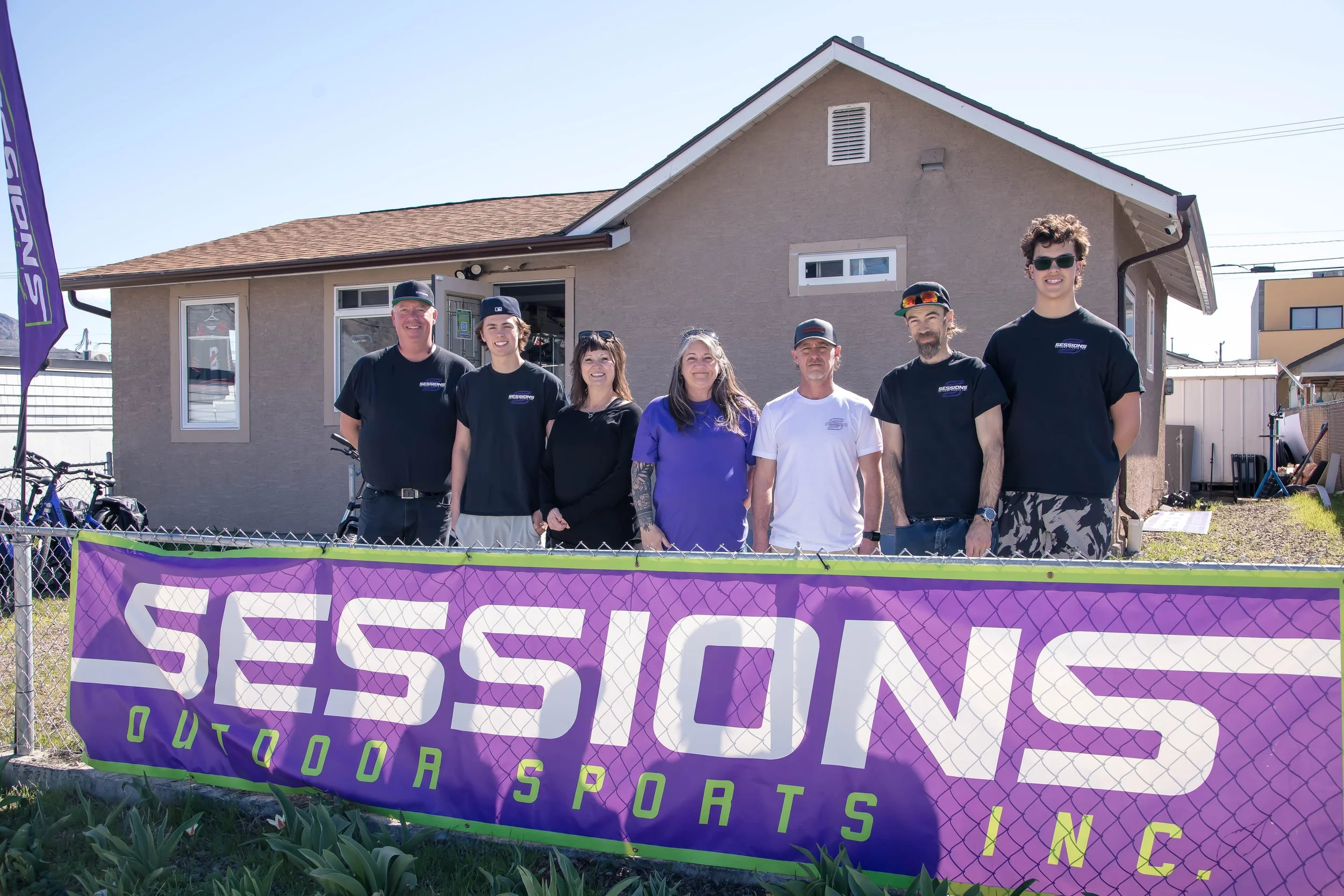 A group of seven people standing behind a purple banner that reads 'SESSIONS OUTDOOR SPORTS INC.', in front of a beige house on a sunny day.